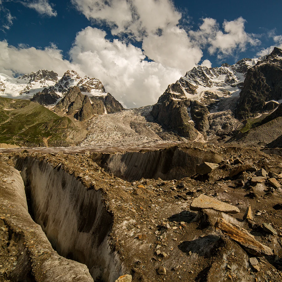 Glacier crevasses in a mountainous landscape with snow-capped peaks under a partly cloudy sky