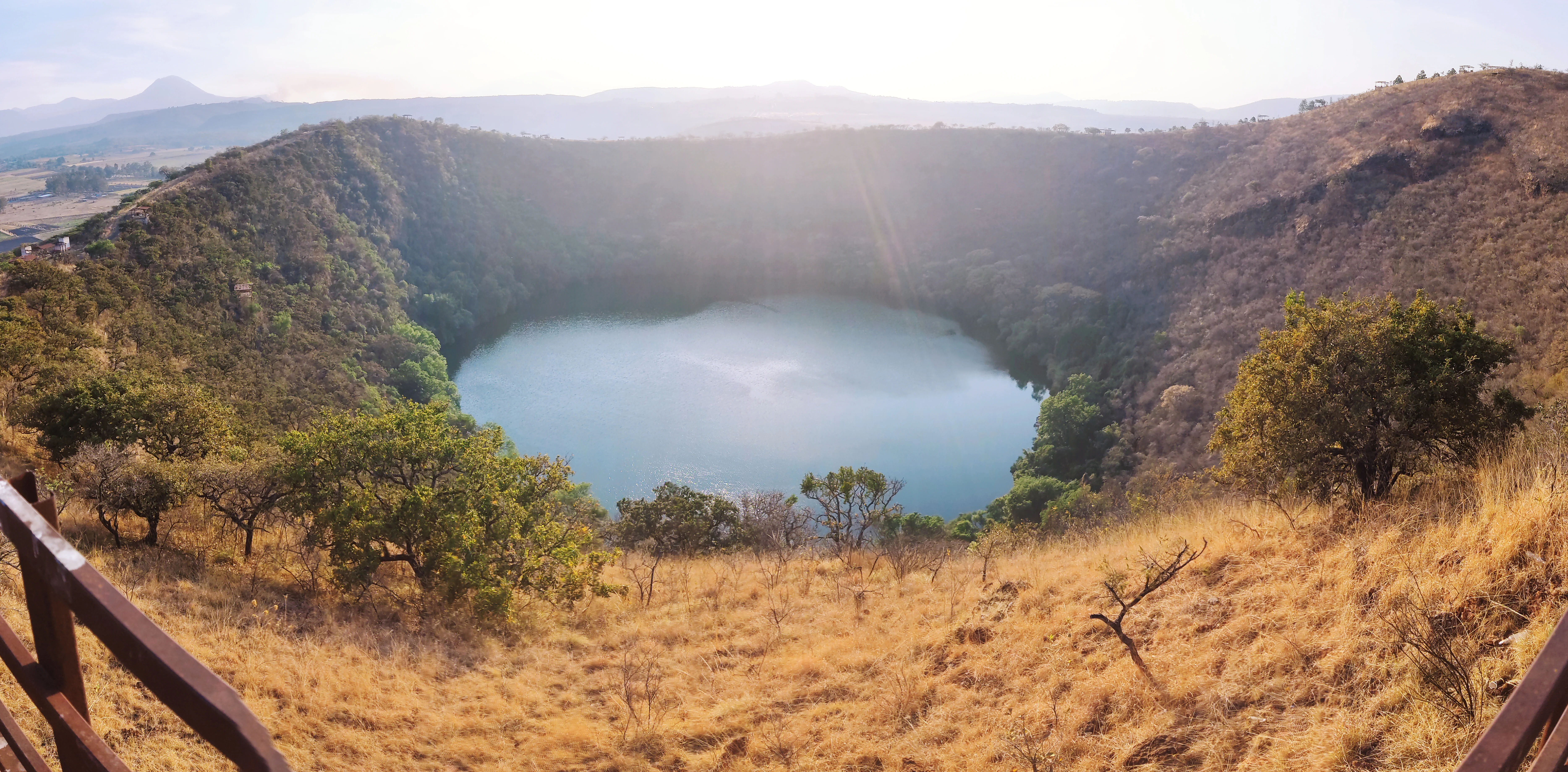 Wide shot of a blue volcanic crater lake surrounded by dry grassy hills and scattered trees, with a wooden fence in the foreground.