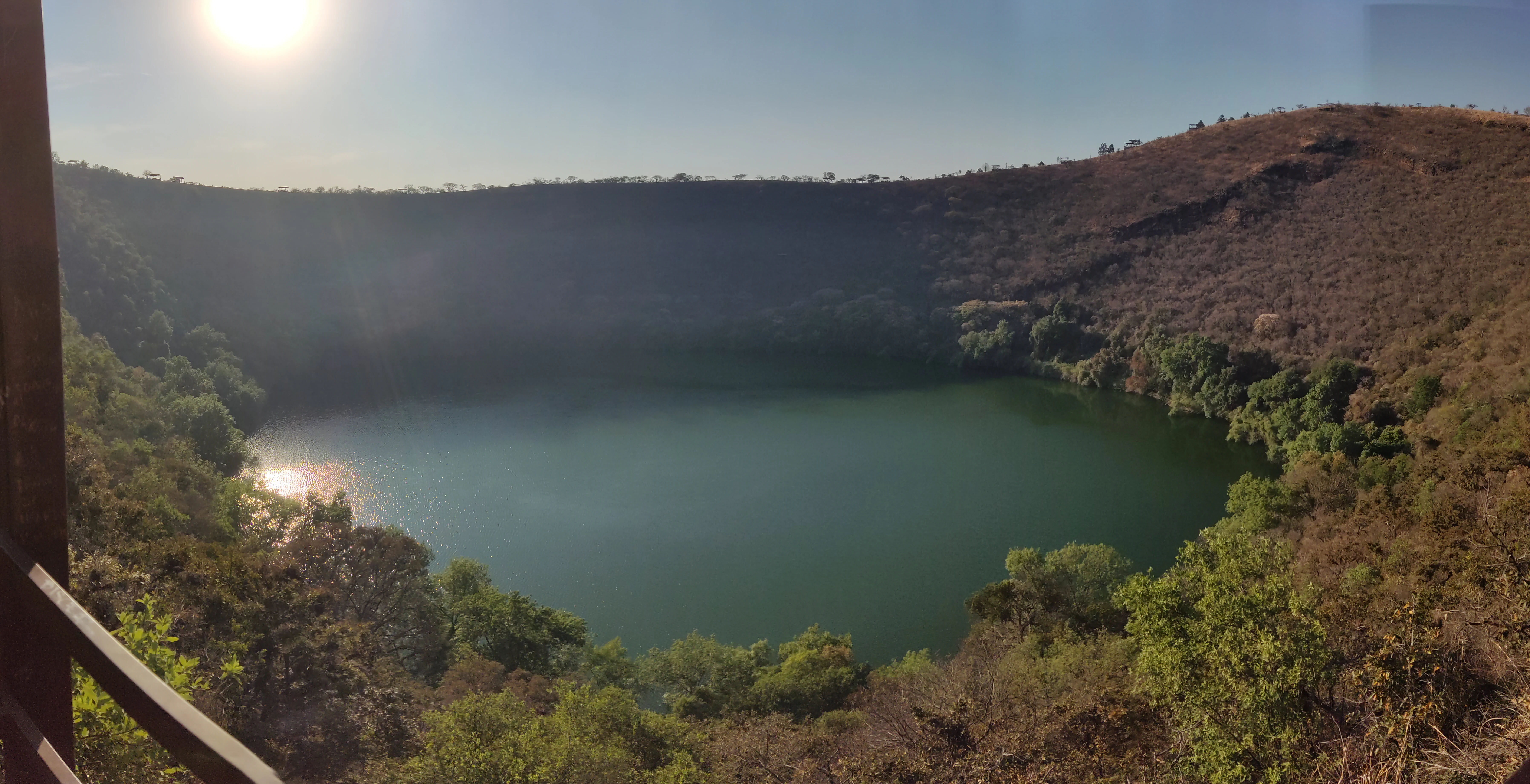 A volcanic crater lake with green water surrounded by steep crater walls covered in dry brown vegetation and scattered green trees, viewed from an elevated wooden viewpoint with the sun shining brightly in the upper left corner.
