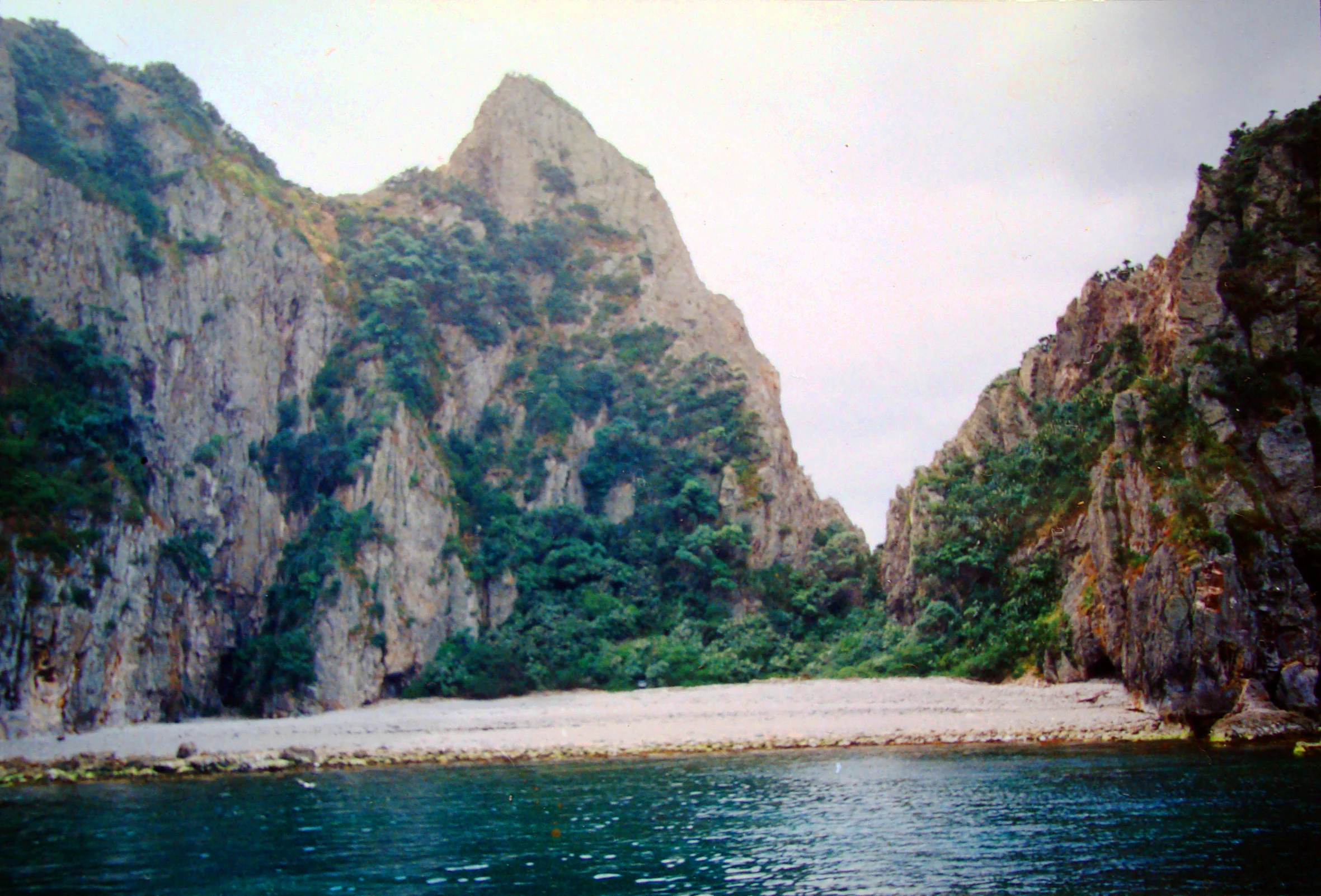 A view from the water looking at a sandy beach nestled between steep, rocky cliffs covered in green vegetation, with a prominent peak in the center.