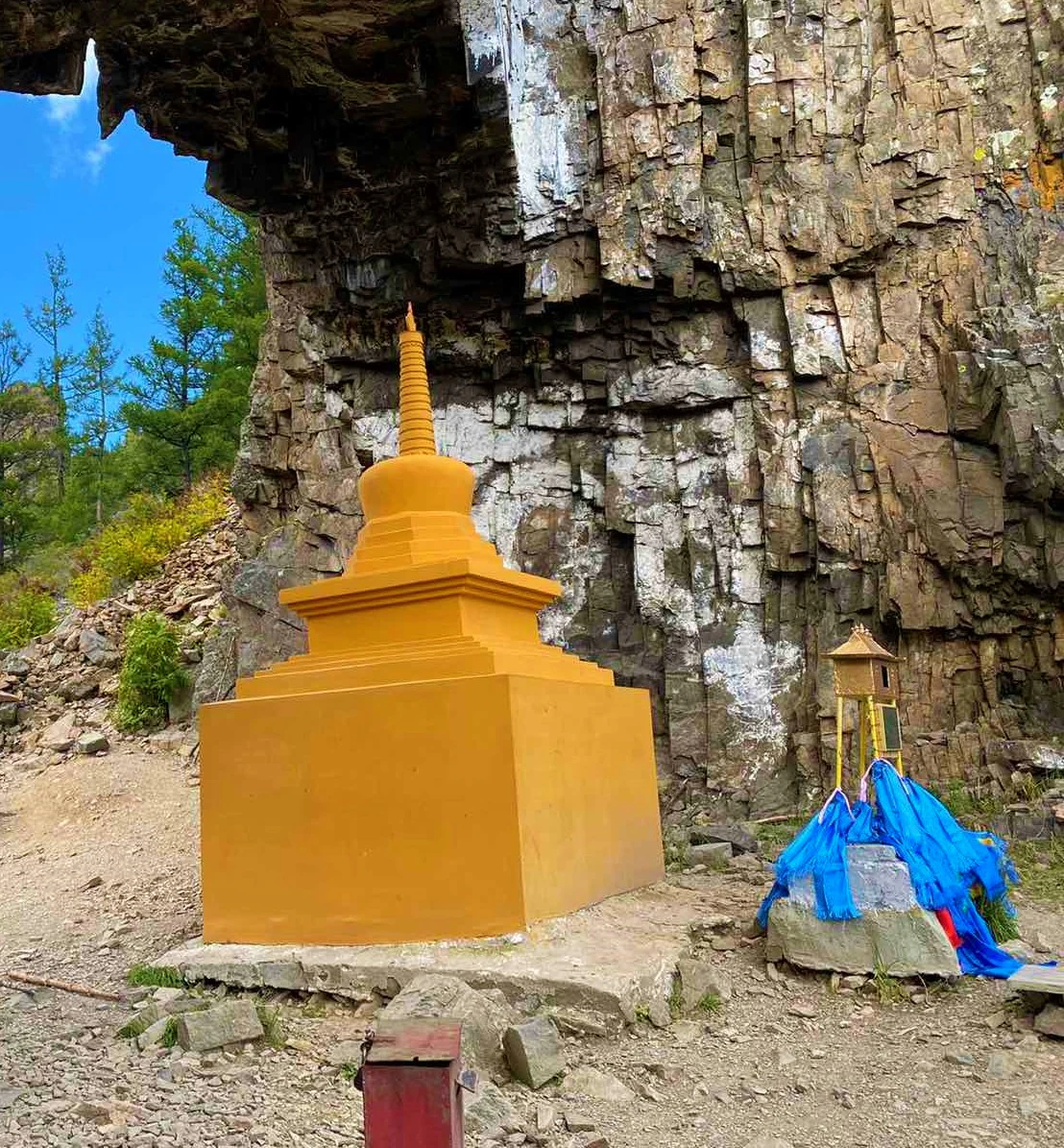 Un suburgán budista de color amarillo brillante se alza de manera prominente sobre un sendero de tierra frente a un acantilado alto y gris de roca estratificada, con paños de oración azules colgados sobre una piedra cercana.