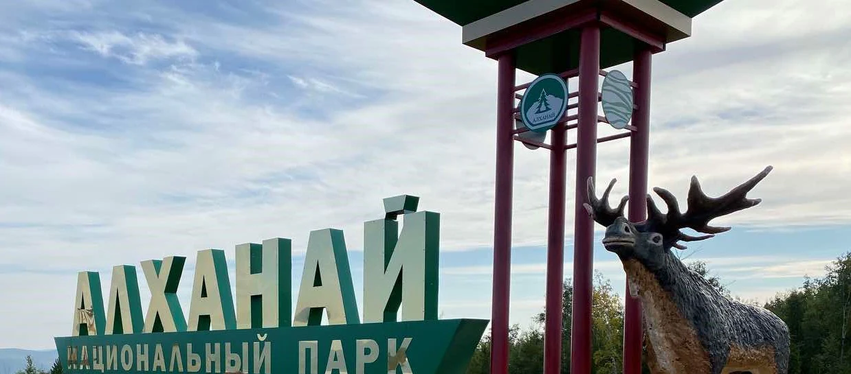 Un gran cartel verde con el texto en ruso del Parque Nacional Alkhanay se encuentra junto a una escultura de madera de un ciervo con grandes astas.