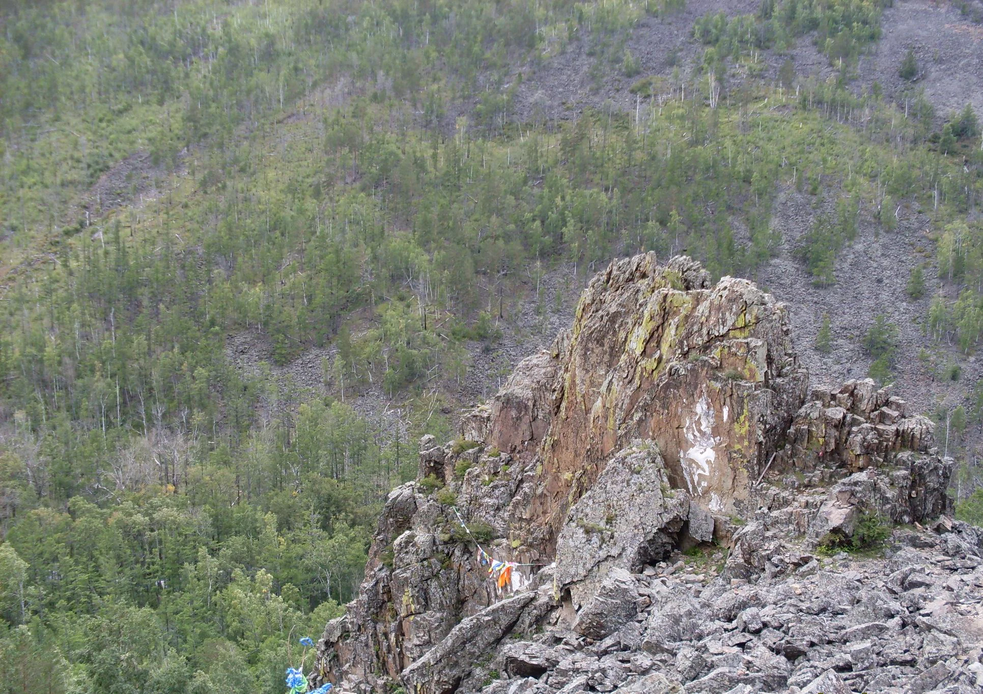 Una formación rocosa vertical y escarpada, conocida como Temple Gate, se alza en medio de una ladera boscosa del Parque Nacional Alkhanay, con banderas de oración atadas cerca de la base.