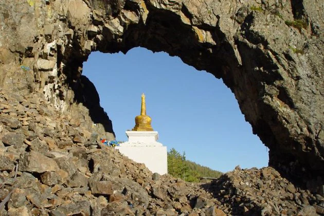 Natural rock arch with a white stupa featuring a golden spire visible through it against a clear blue sky.