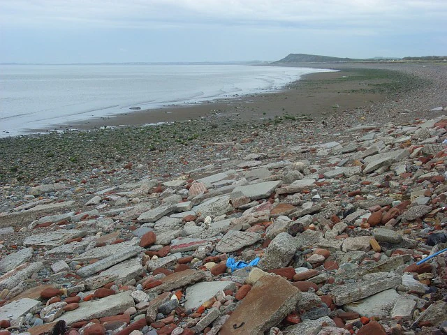 A wide angle view of a stony and debris-filled shoreline meeting the calm waters of the Solway Firth under a cloudy sky.