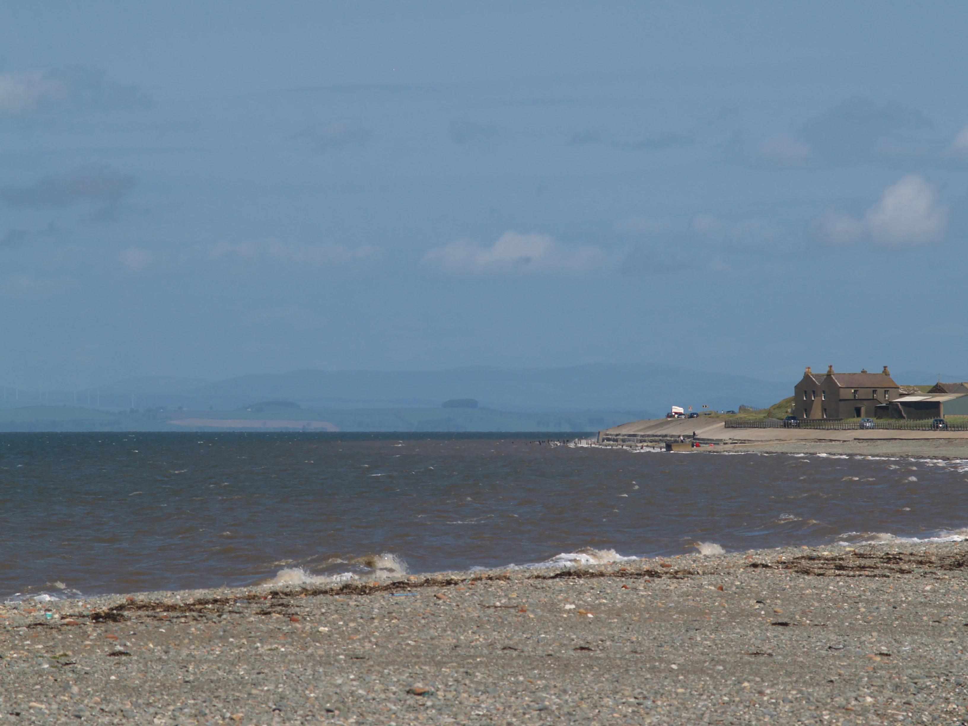 A wide shot of a gravel beach meeting the calm waters of the Solway Firth, with buildings visible on the shoreline to the right and distant blue hills across the bay.