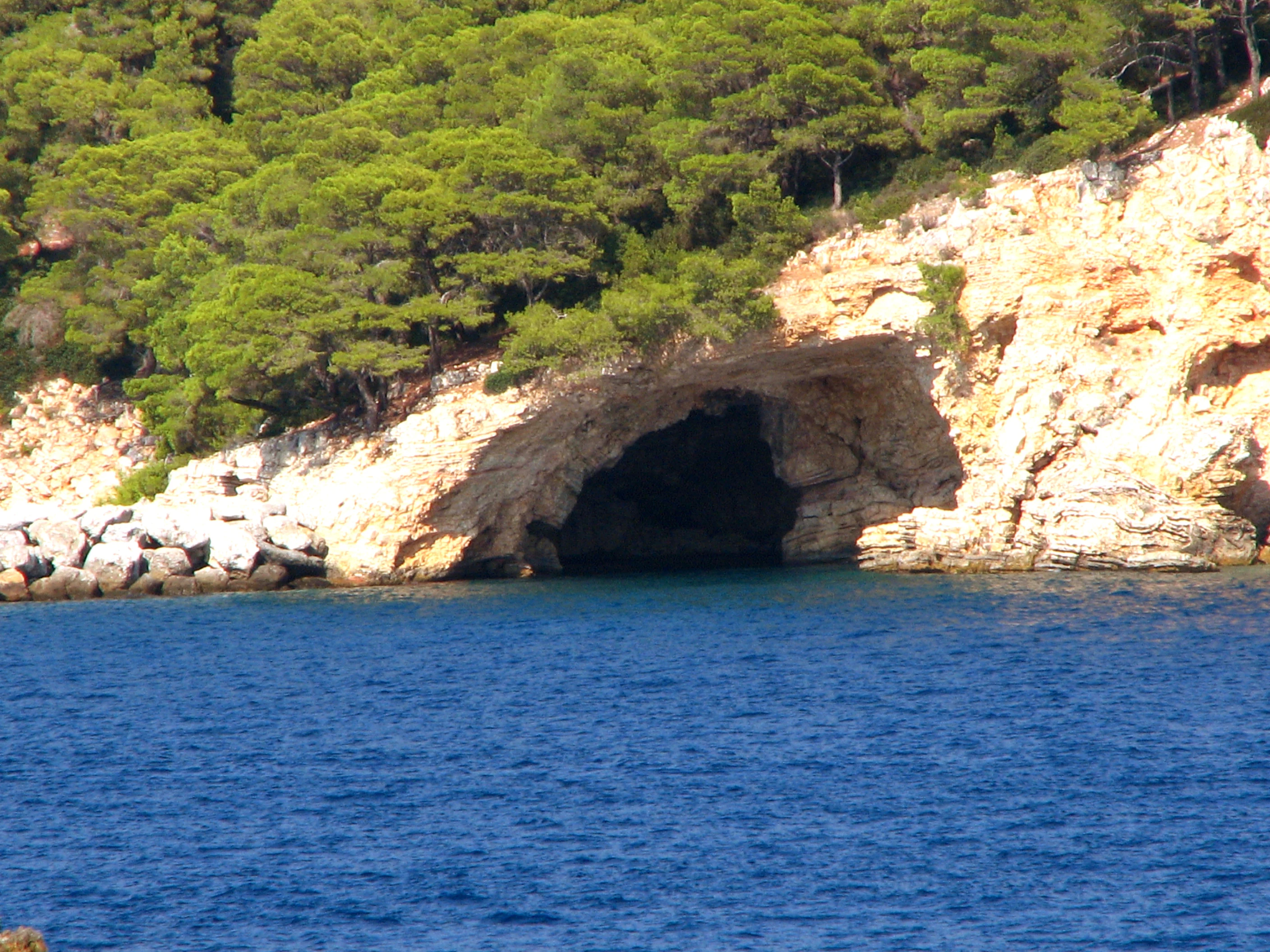 Blue sea in foreground, rocky cliffs with cave opening, green trees in background.