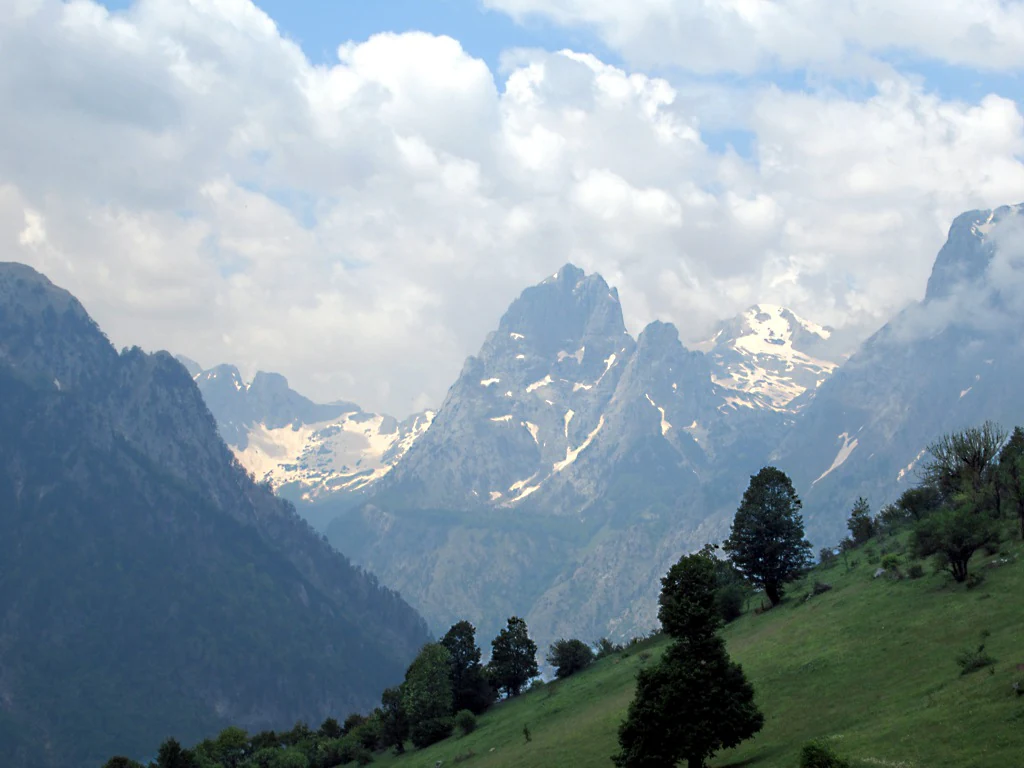 Wide view of mountain peaks with patches of snow, green valleys, and scattered trees under a partly cloudy sky