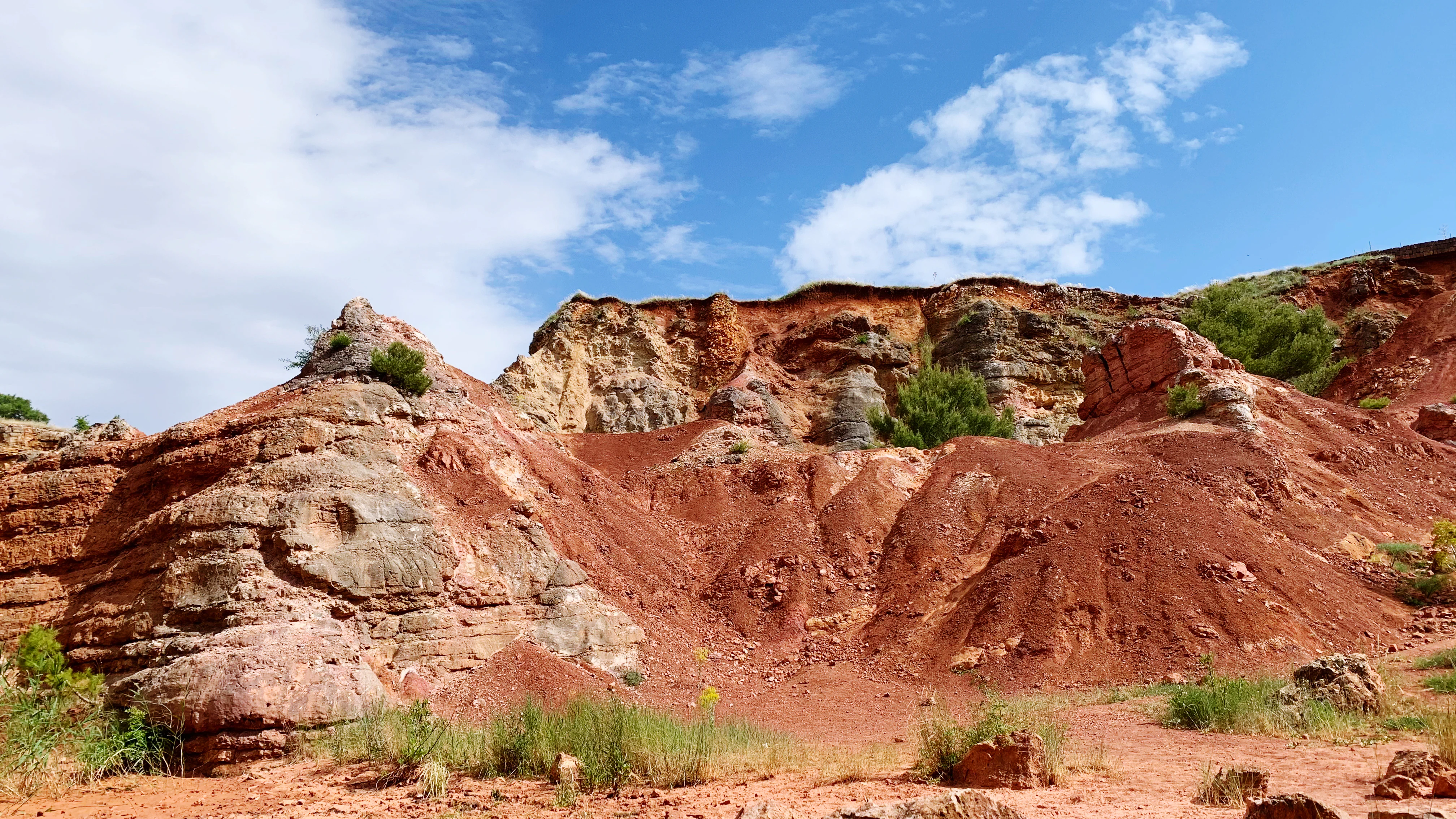 Red and brown rock formations with sparse vegetation under blue sky with scattered clouds