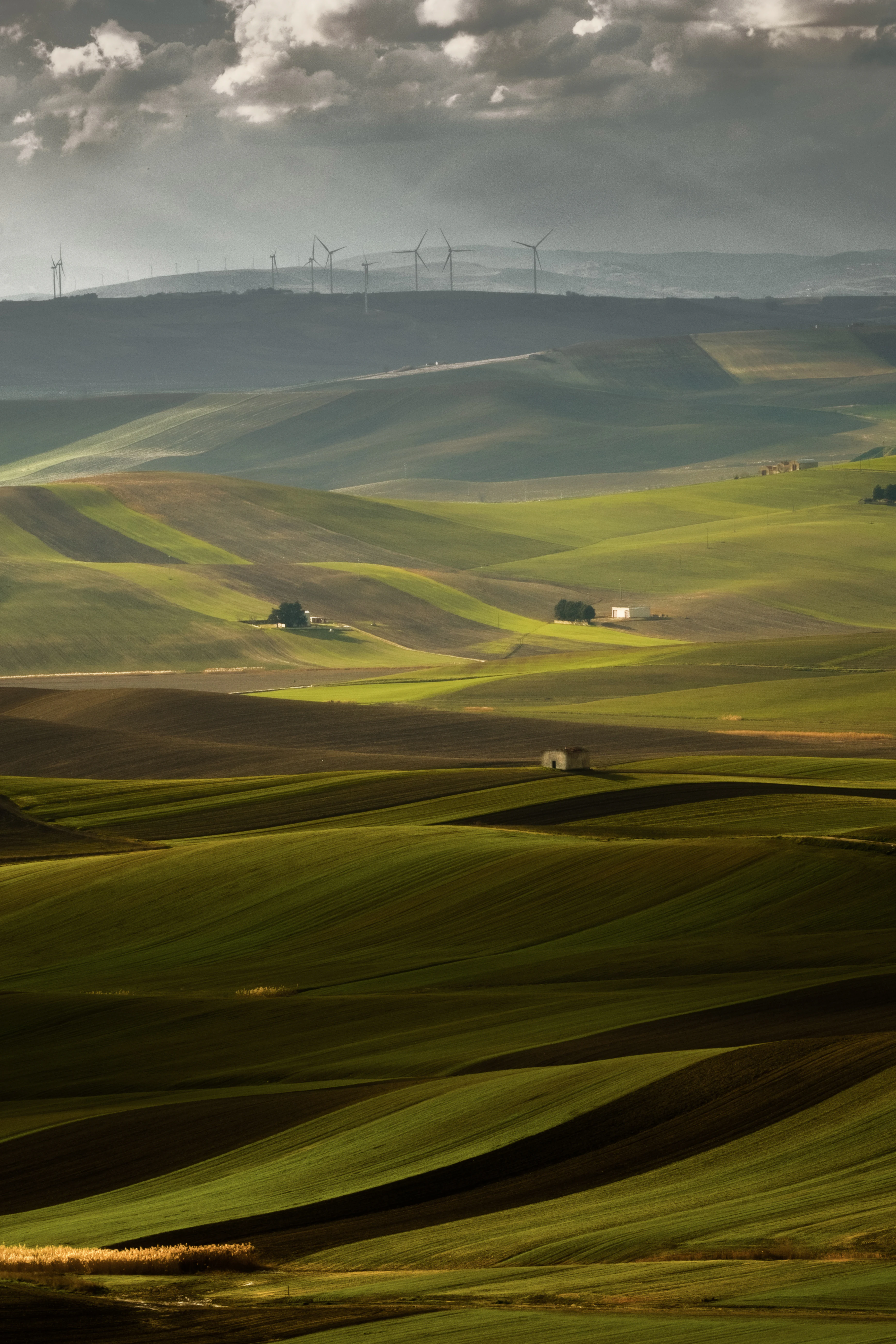 Wide landscape view of rolling green hills with small structures and distant wind turbines under a partly cloudy sky