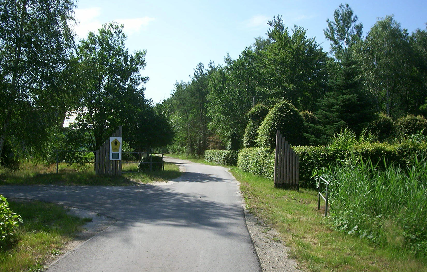 A paved path leads through a park entrance marked by two wooden posts and a nature reserve sign, bordered by trees and dense green vegetation.