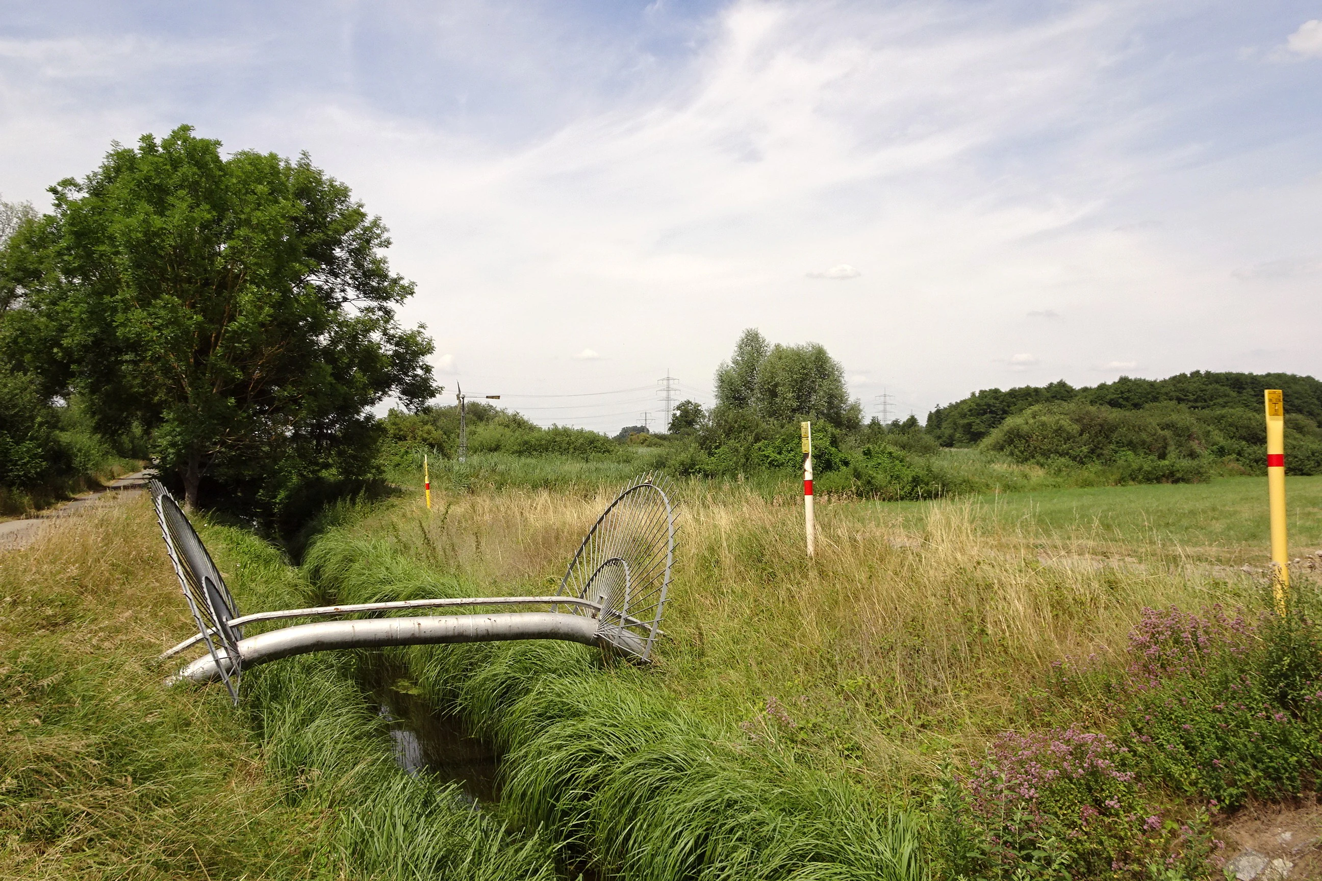 A landscape view of a rural nature reserve with a small drainage ditch in the foreground, yellow pipeline markers, and distant trees under a partly cloudy sky.