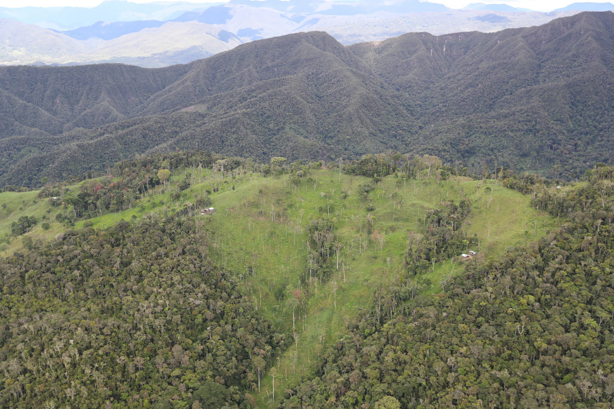 An aerial view showing green tropical forest covering mountain slopes with a clearing or pasture visible in the midground.
