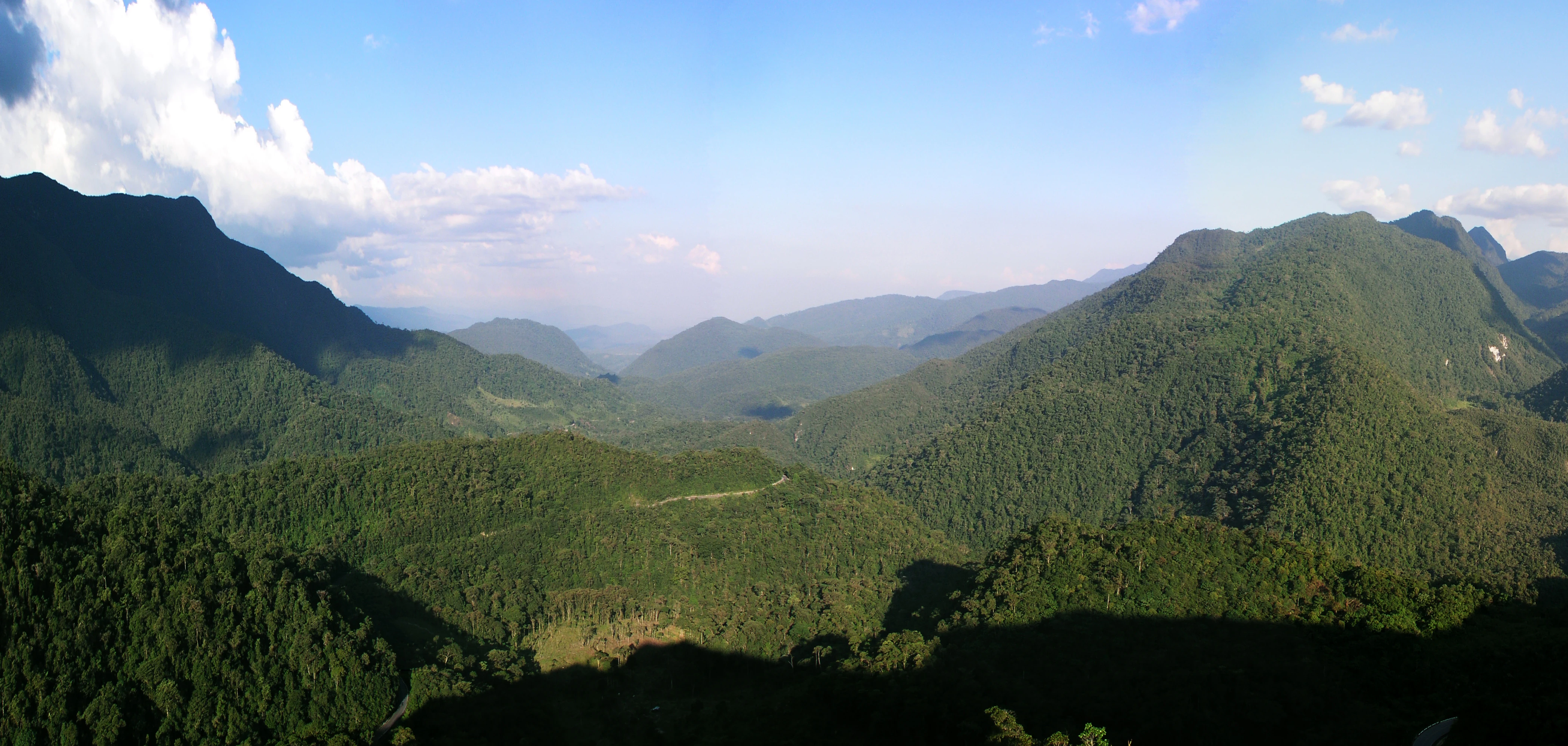 A wide panoramic view of lush, forested mountains under a blue sky with scattered white clouds, taken from a high vantage point.