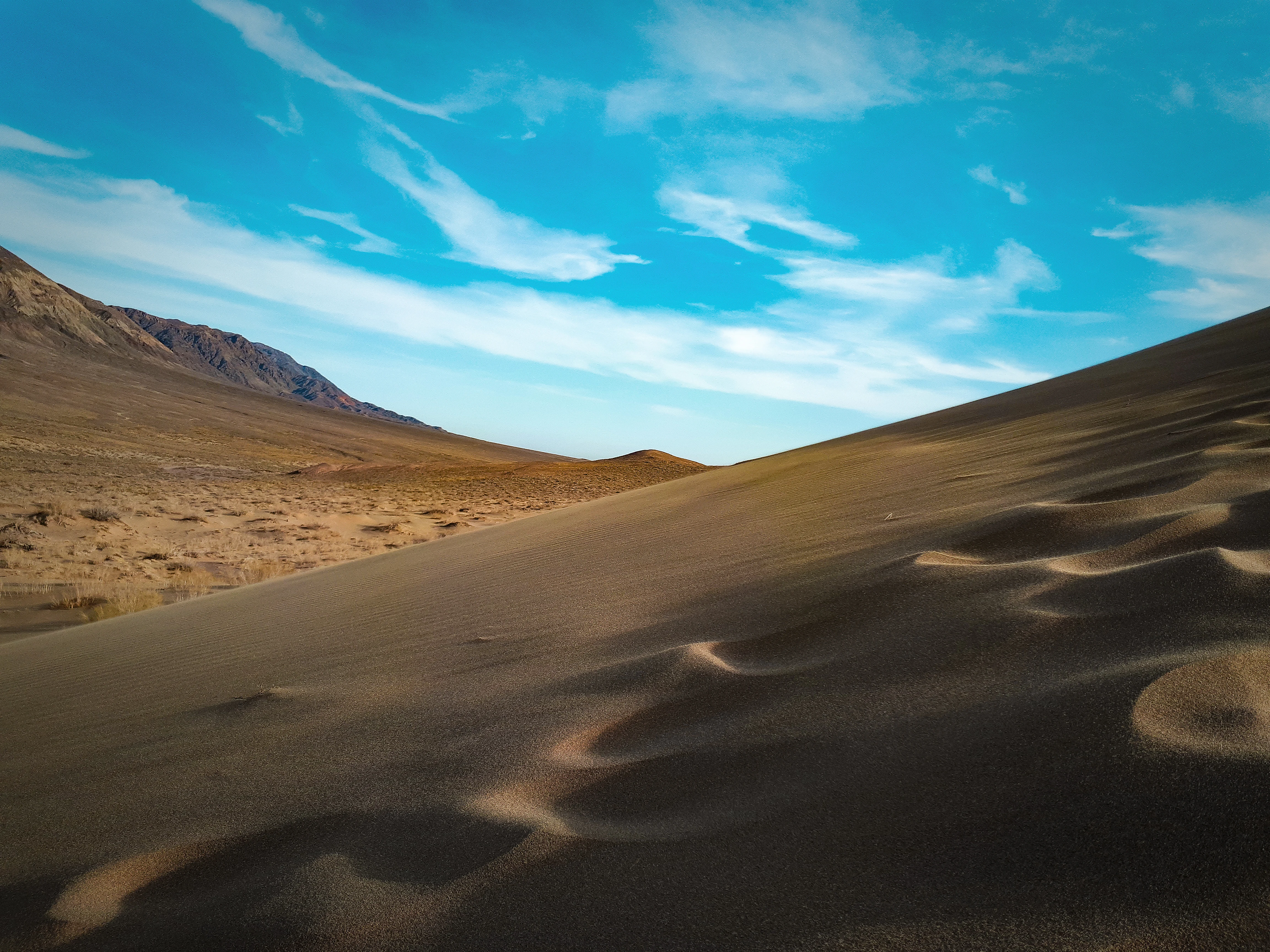 Sand dunes with visible wind patterns under a clear blue sky with scattered clouds