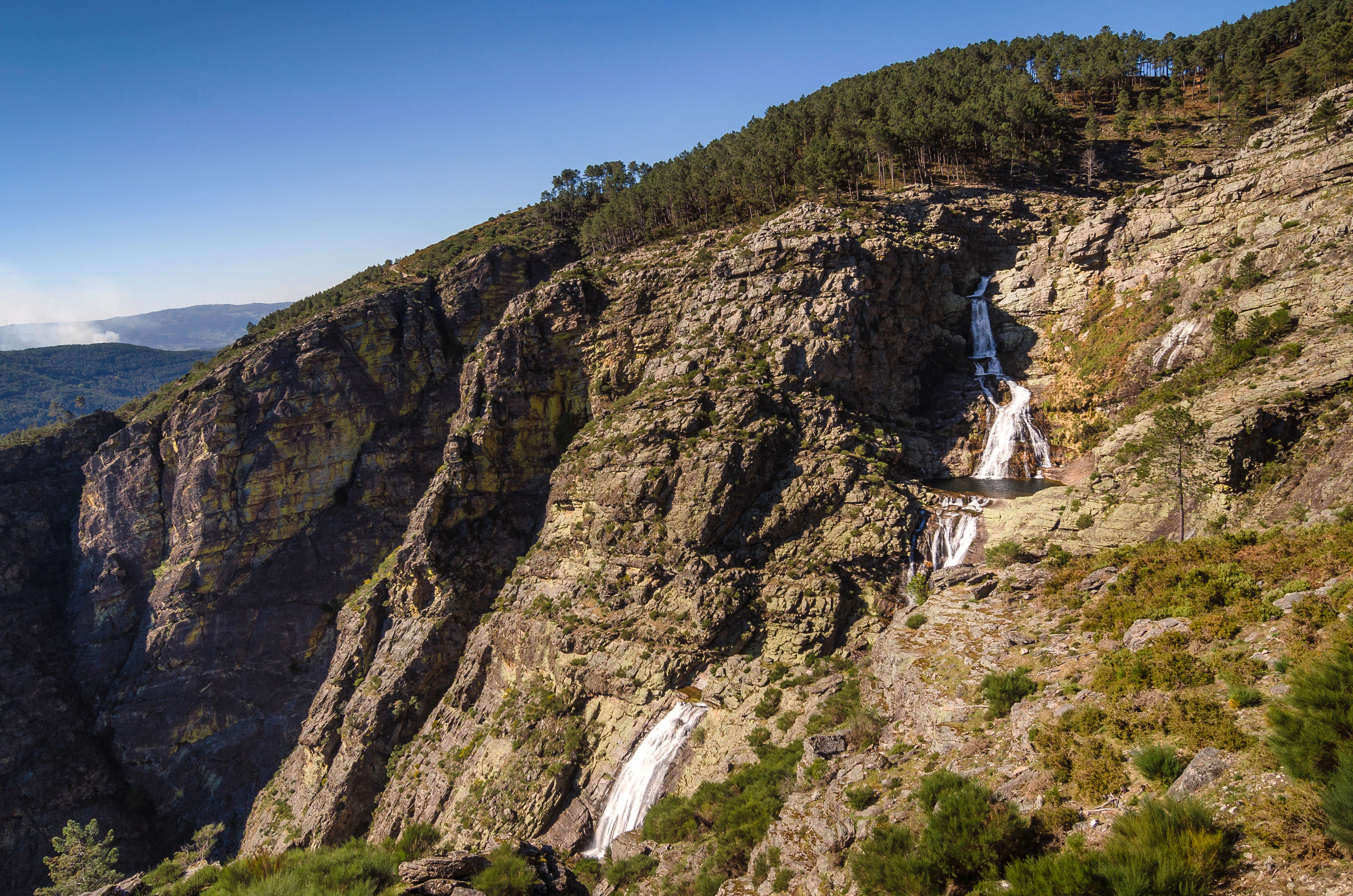 A waterfall cascading down a rocky cliff face, surrounded by sparse vegetation and trees, under a clear blue sky