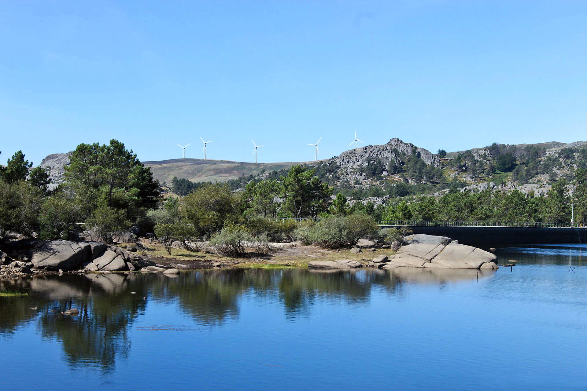 Reservoir with clear blue water reflecting rocks, trees, and hills under a clear sky