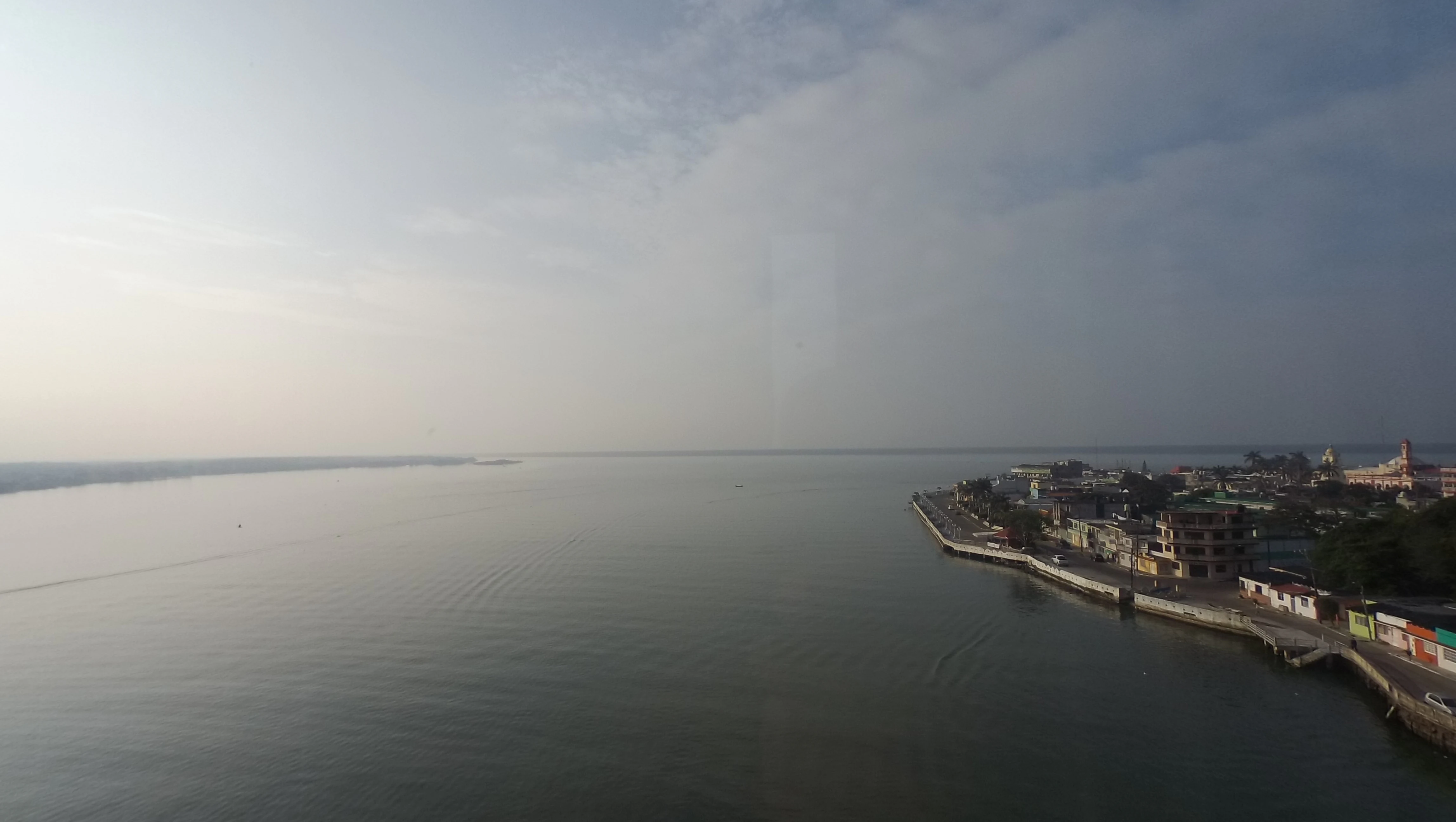 An elevated wide shot looking out over a calm, expansive body of water bordering the town of Alvarado, featuring a coastal promenade and urban buildings along the shoreline.