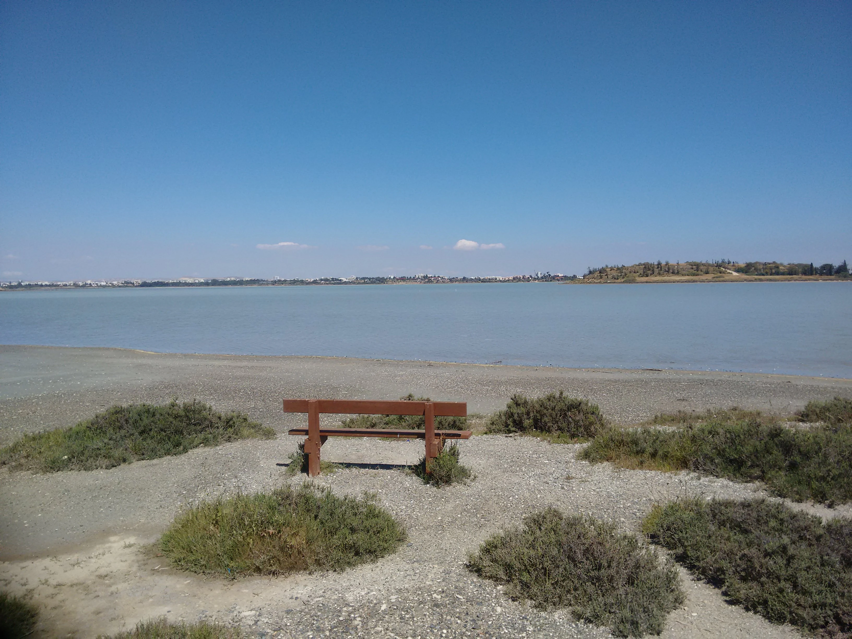 A brown wooden bench sitting on a gravelly, shrub-covered shore facing a calm, wide salt lake under a bright blue sky.
