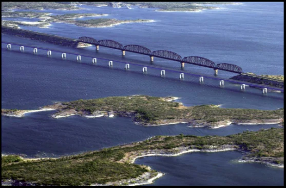An aerial view showing two long bridges crossing a large body of dark blue water with several small, rocky islands and shrubby vegetation in the foreground.