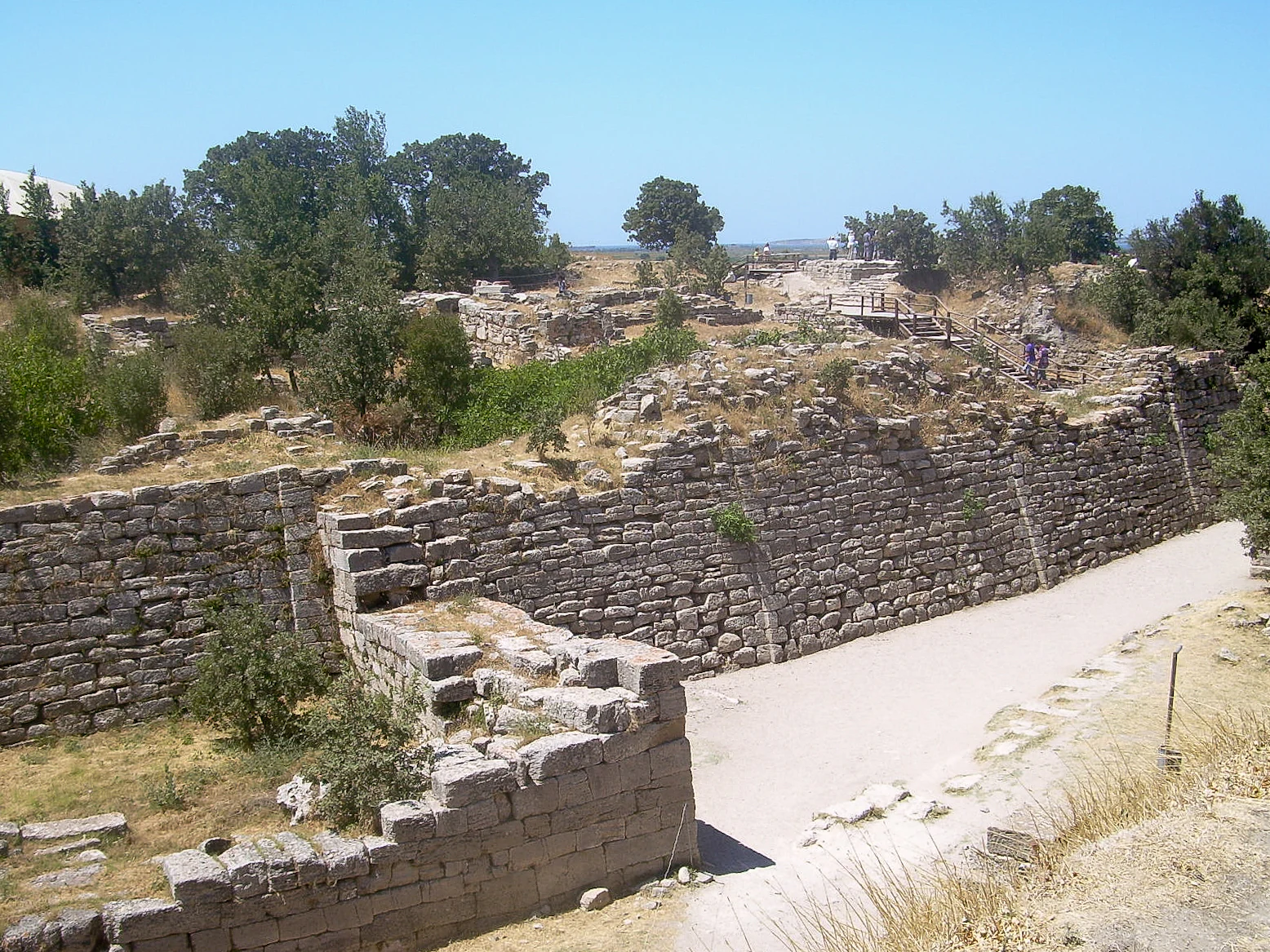 Ancient stone walls of Troya National Park with stone pathway, dry grass, and scattered trees under clear blue sky
