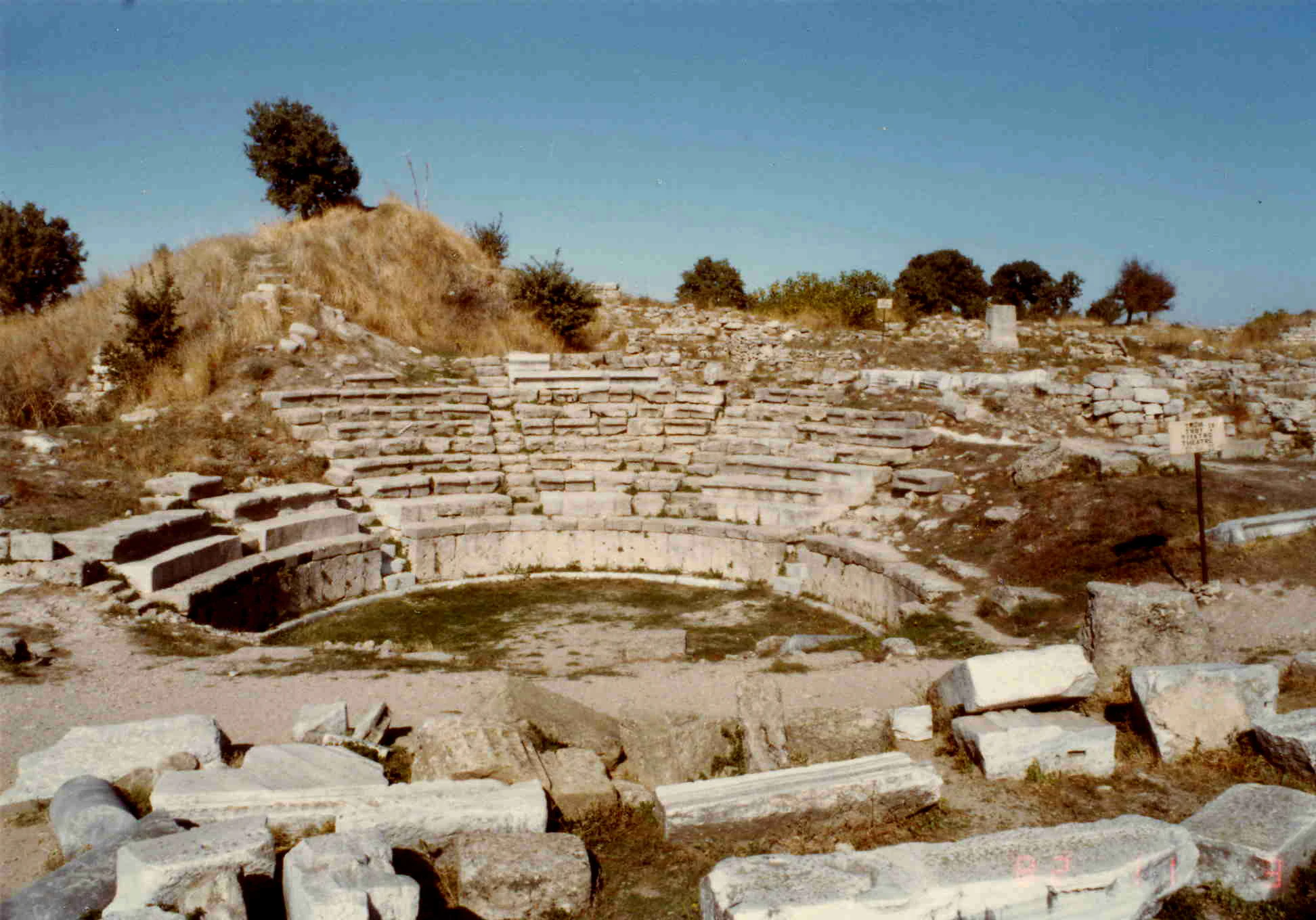 Stone amphitheater ruins with tiered seating, grassy center, and scattered stone blocks under clear sky