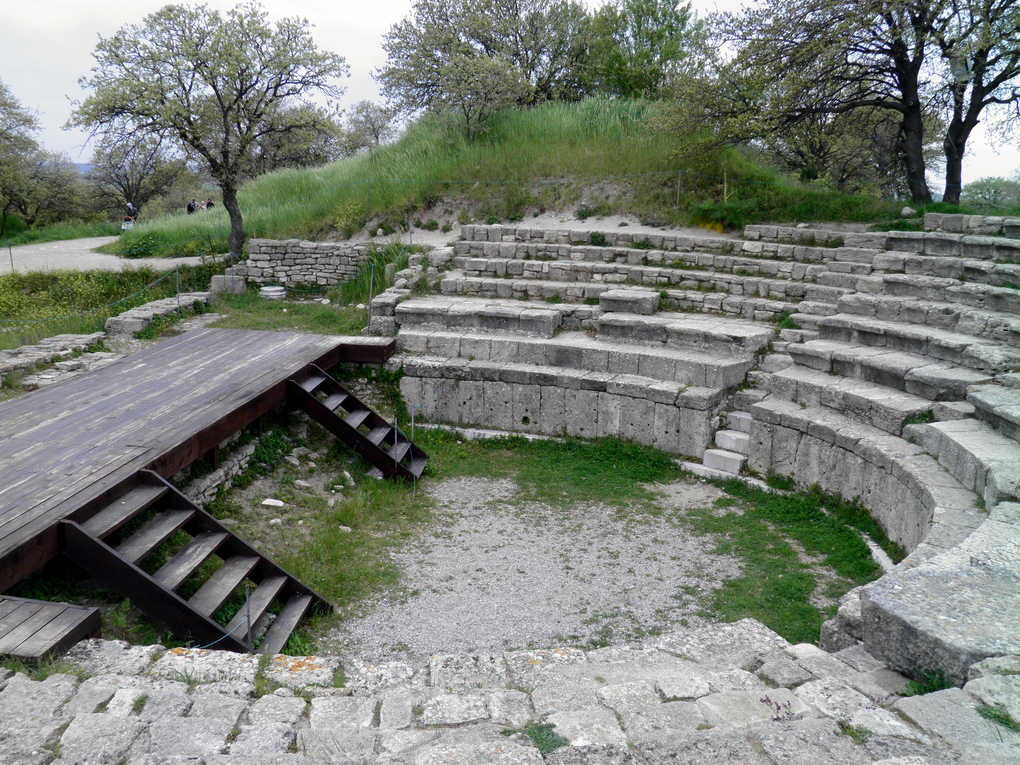 Stone amphitheater with tiered seating, wooden platform with stairs, grassy area, and trees in the background