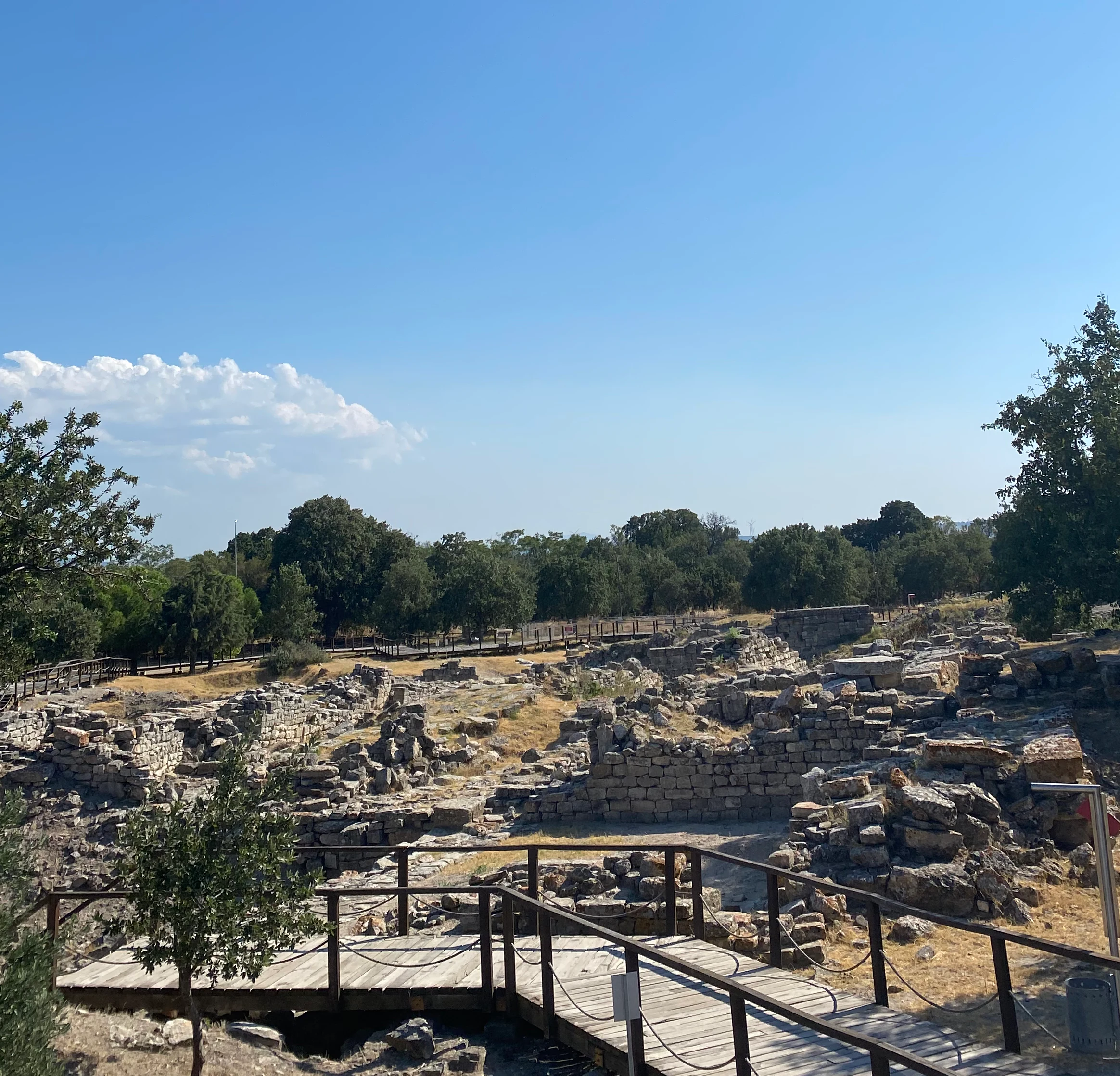 Ancient stone ruins with wooden walkway and railings, surrounded by trees under a clear blue sky with scattered clouds.