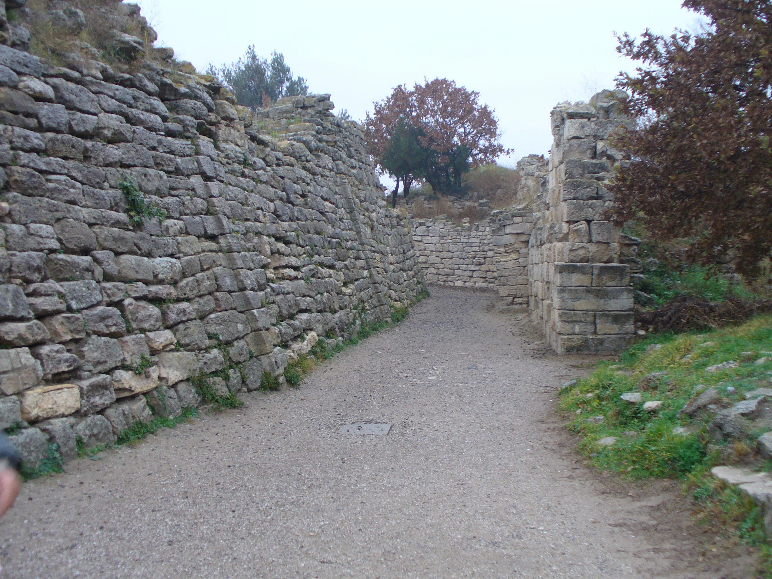 Stone walls forming an ancient pathway with a gravel path between them, trees in the background