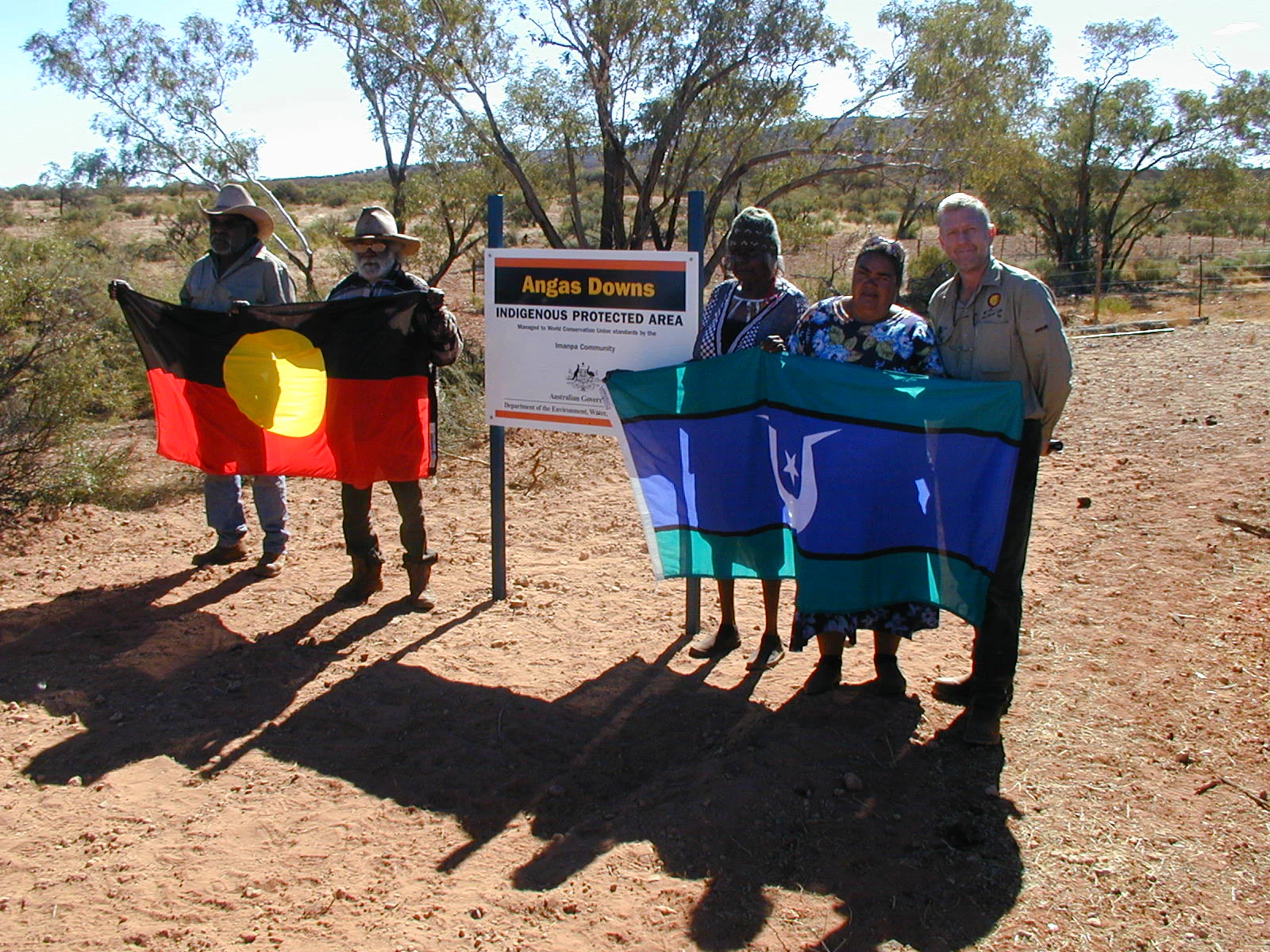 A group of five people stand in an arid outdoor setting, holding flags beside a formal sign that reads Angas Downs Indigenous Protected Area.