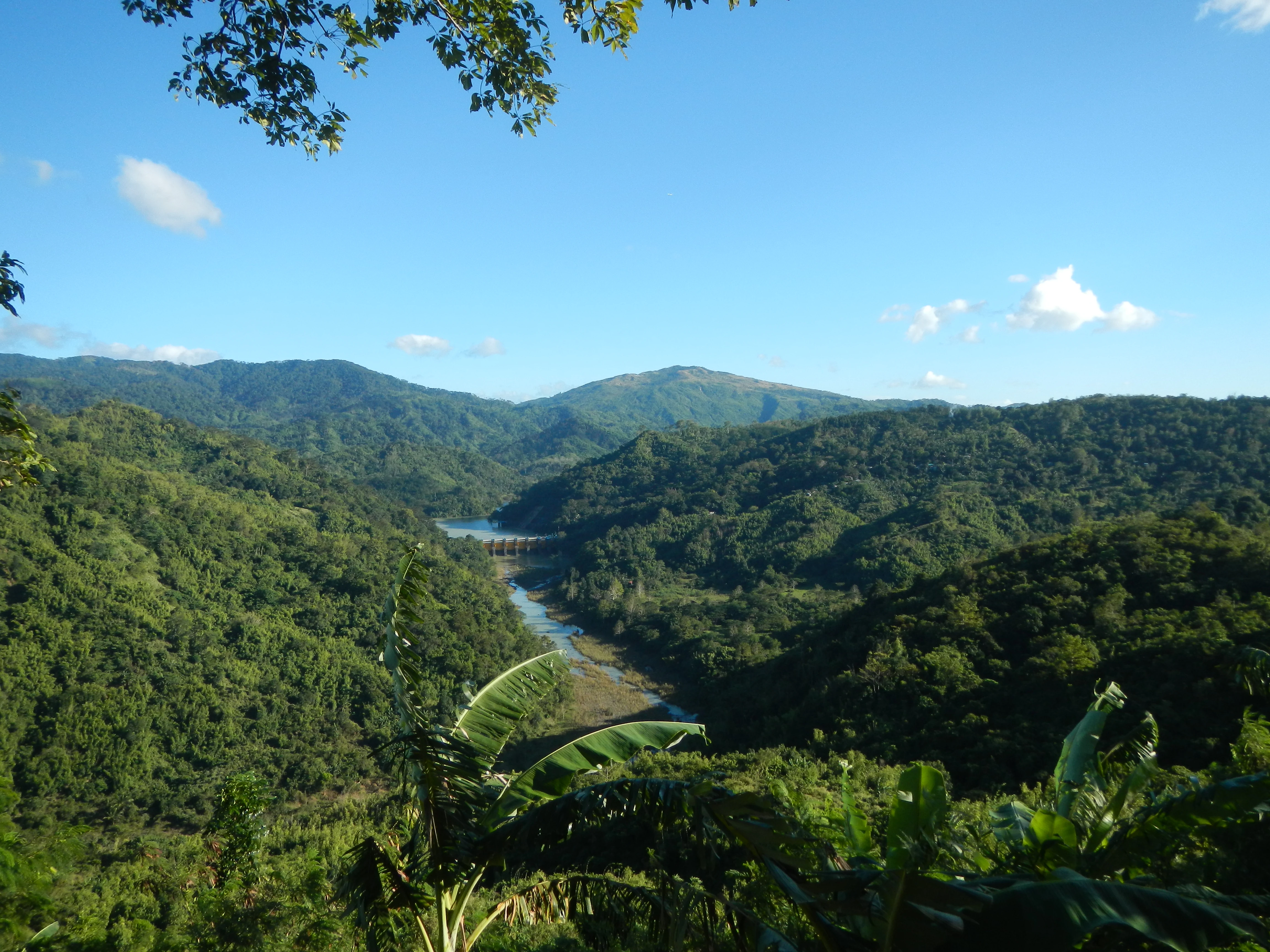 A wide, high-angle view of a river valley containing a dam structure, surrounded by dense, rolling green hills and mountains under a clear blue sky.
