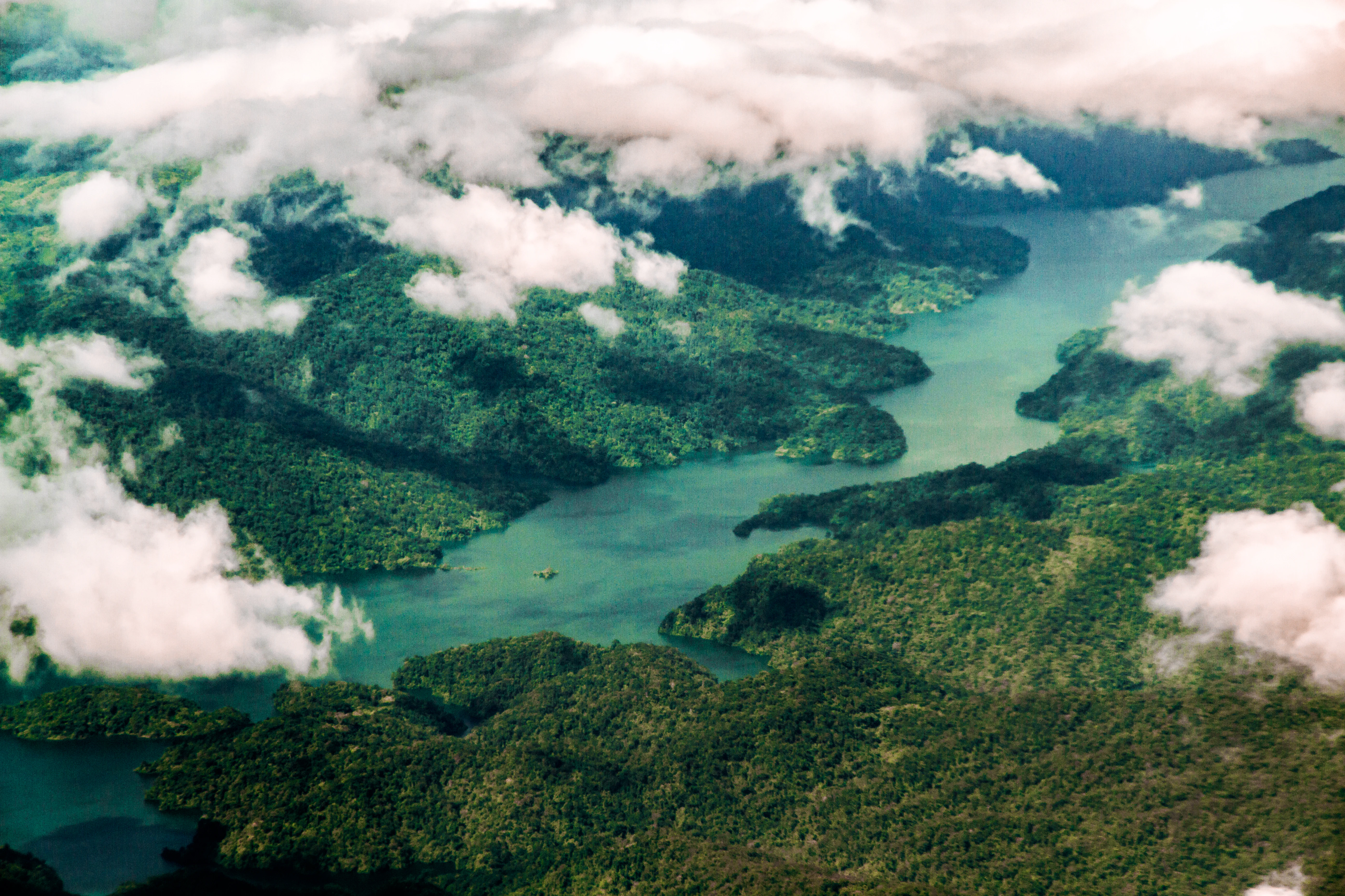 Aerial view looking down at a large, winding reservoir filled with blue-green water surrounded by dense, lush green hills and forests, with scattered white clouds partially obscuring the landscape.