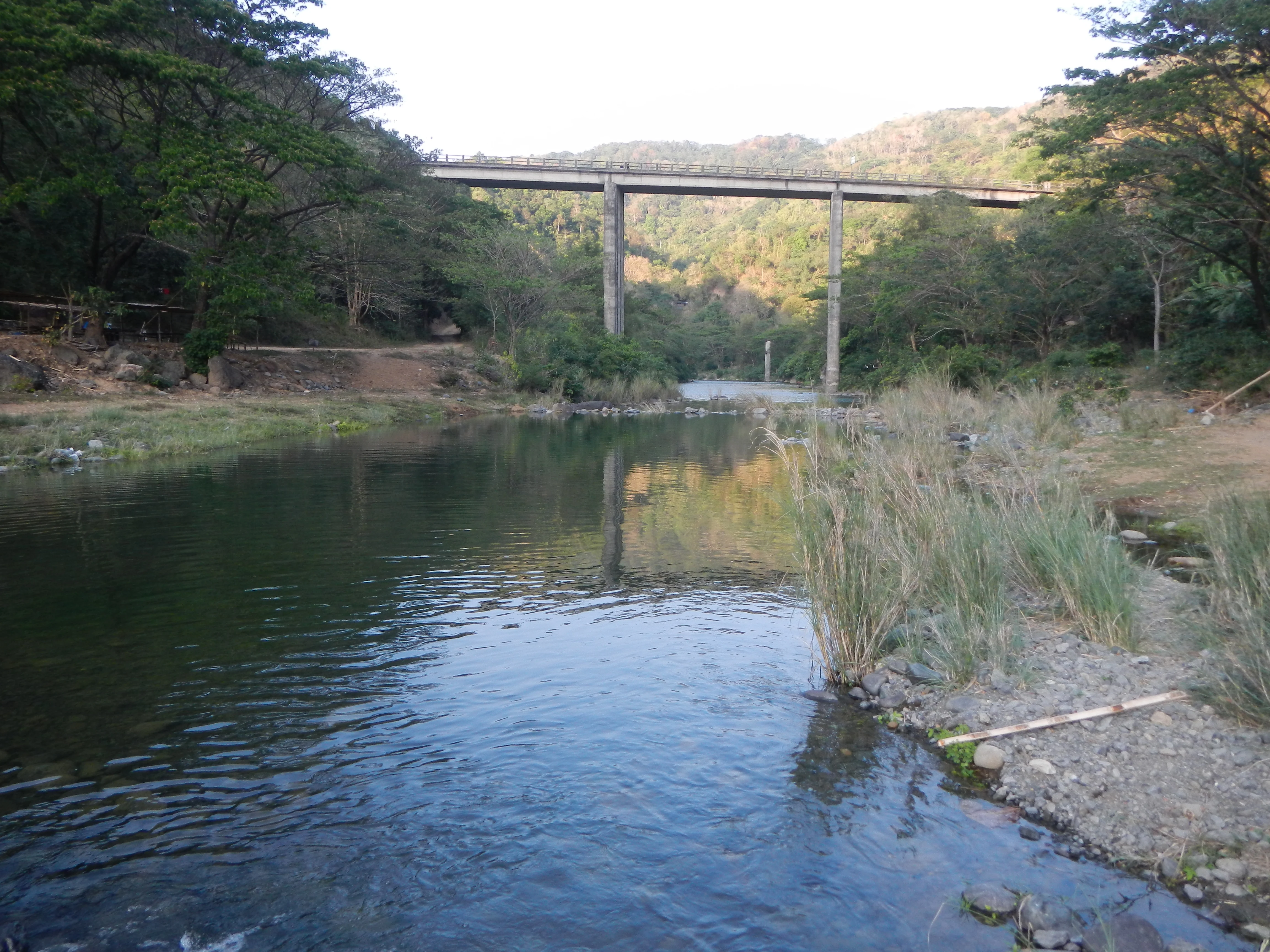A concrete bridge stands above a calm river surrounded by forest and rolling hills in a rural landscape.