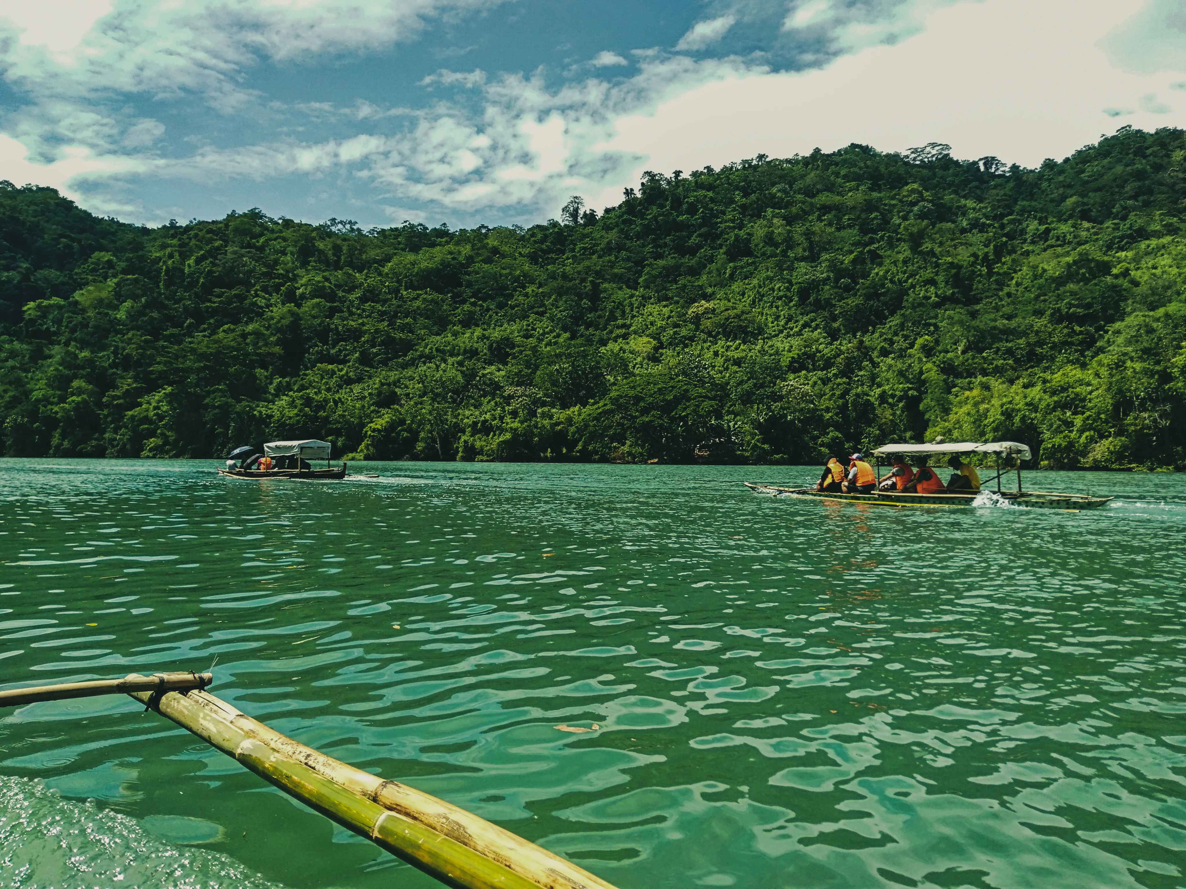 A view from the front of a small boat showing two other passenger boats on a large, green-tinted reservoir surrounded by dense forest-covered hills under a cloudy sky.