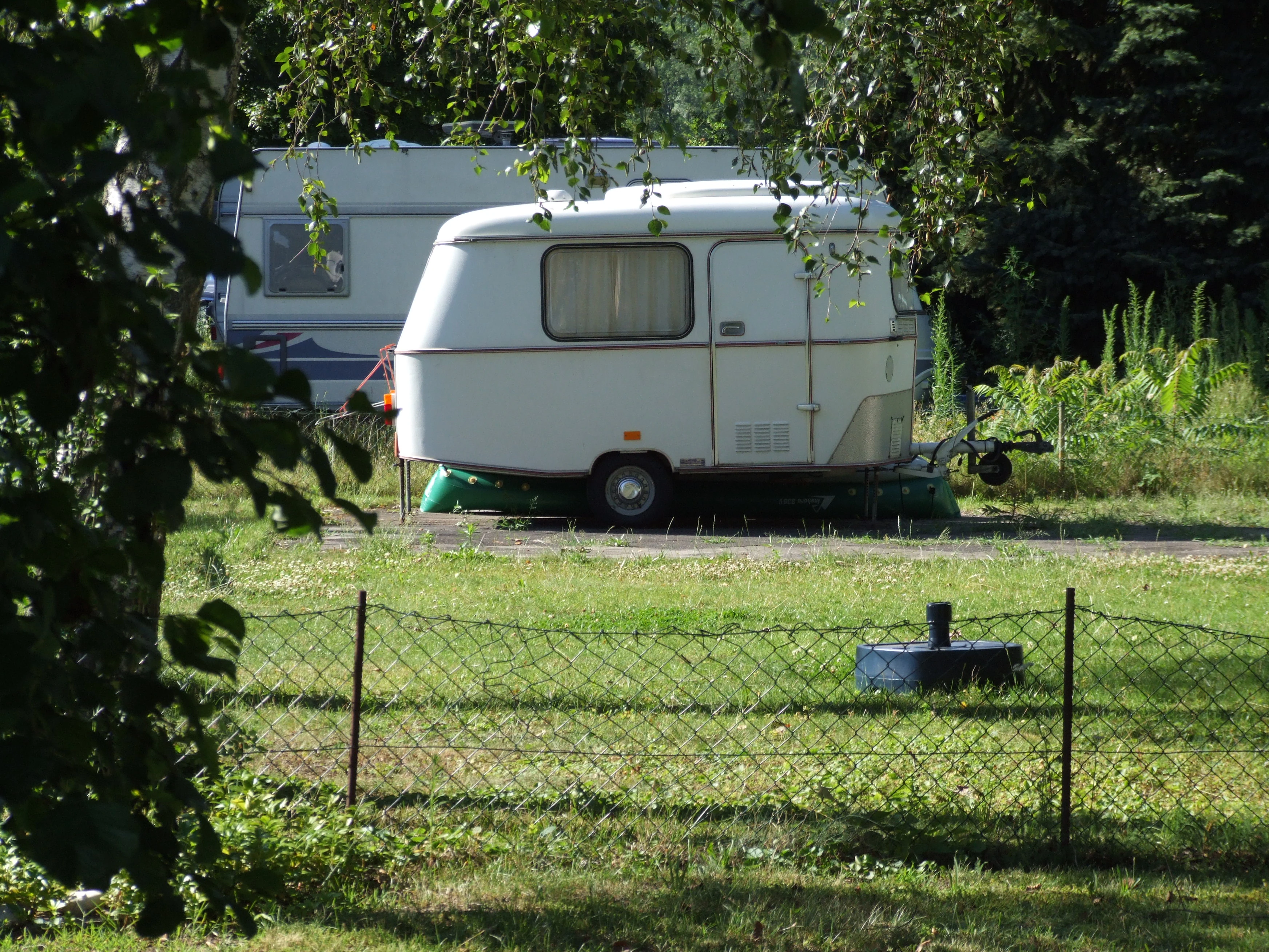 A white vintage camper trailer parked on a grassy field behind a wire fence, with another larger caravan partially visible in the background.