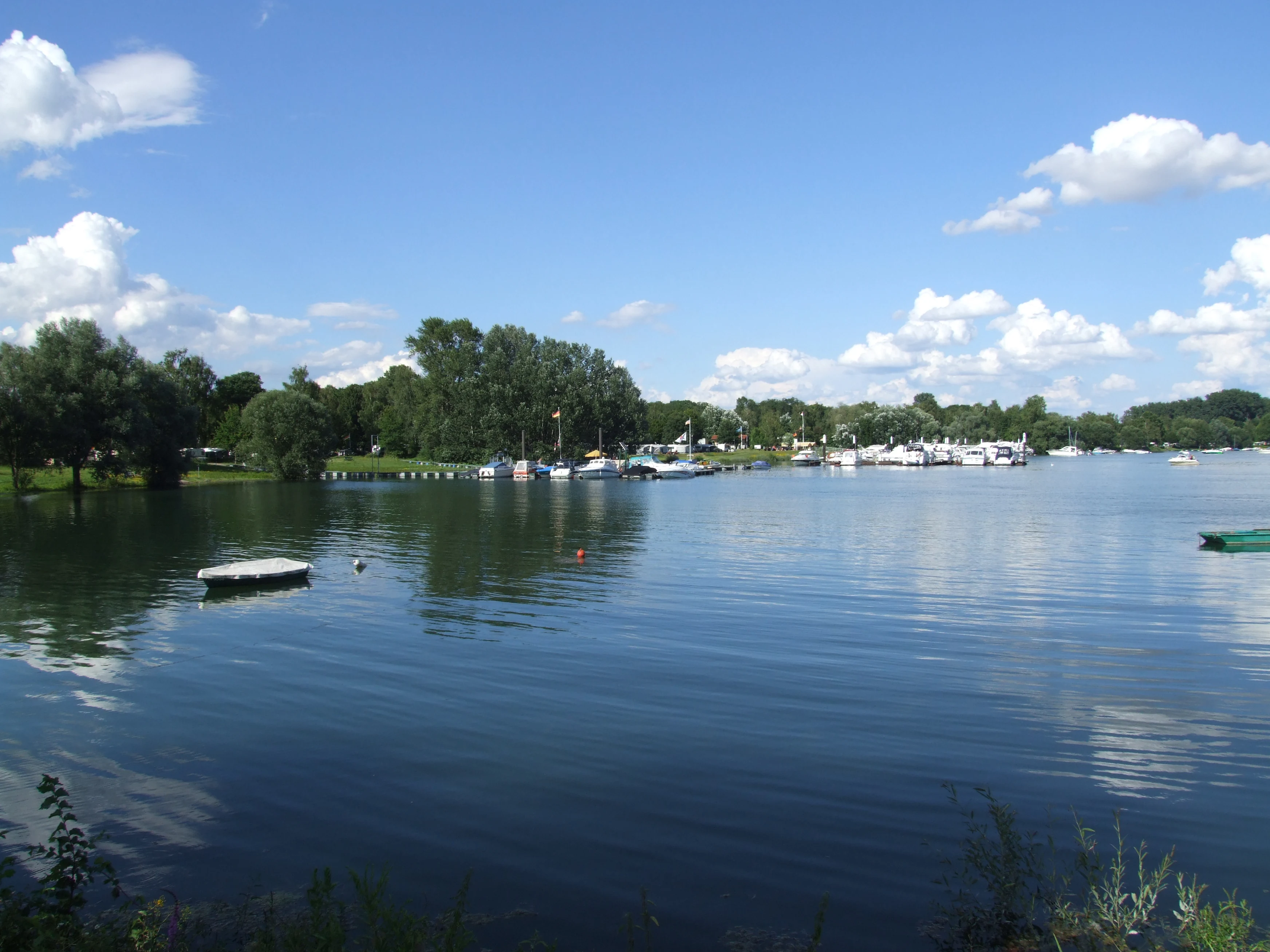 A wide view of the Angelhofer Altrhein waterway with moored boats, green trees on the banks, and a blue sky with scattered clouds.