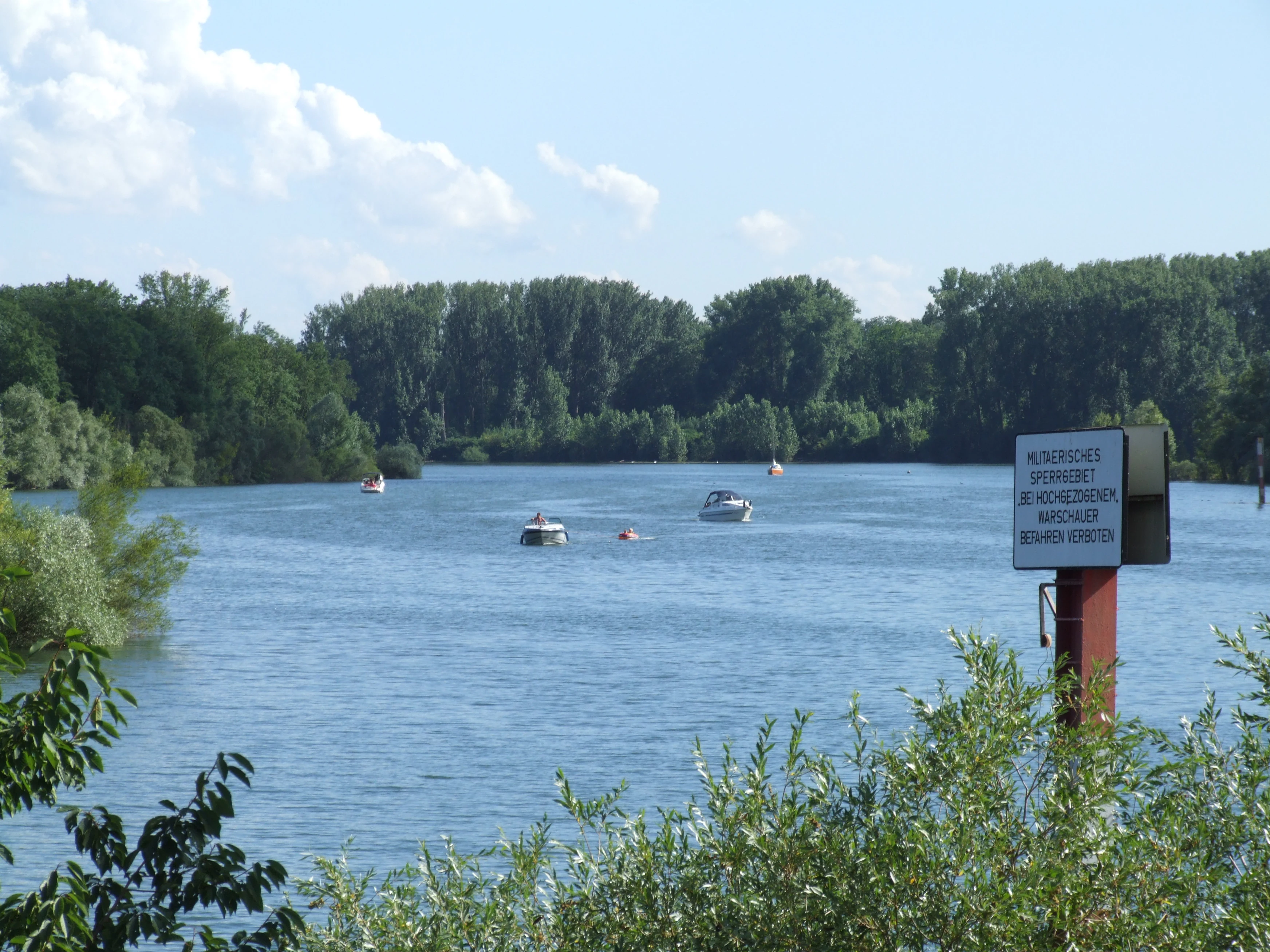 A wide view of a calm, blue river branch, the Angelhofer Altrhein, with several small boats on the water and a square information sign on a post in the right foreground, set against a backdrop of dense green trees.