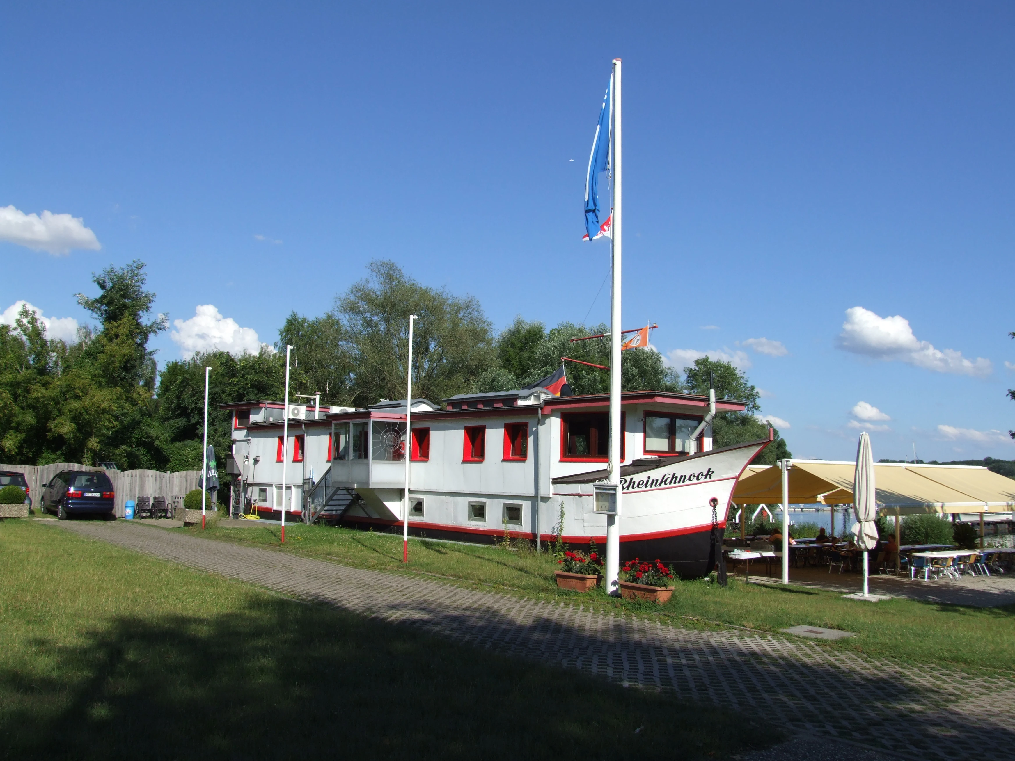 A white building shaped like a ship with red trim, serving as a clubhouse, situated on a grassy area next to a paved path and a tent-like outdoor seating area.
