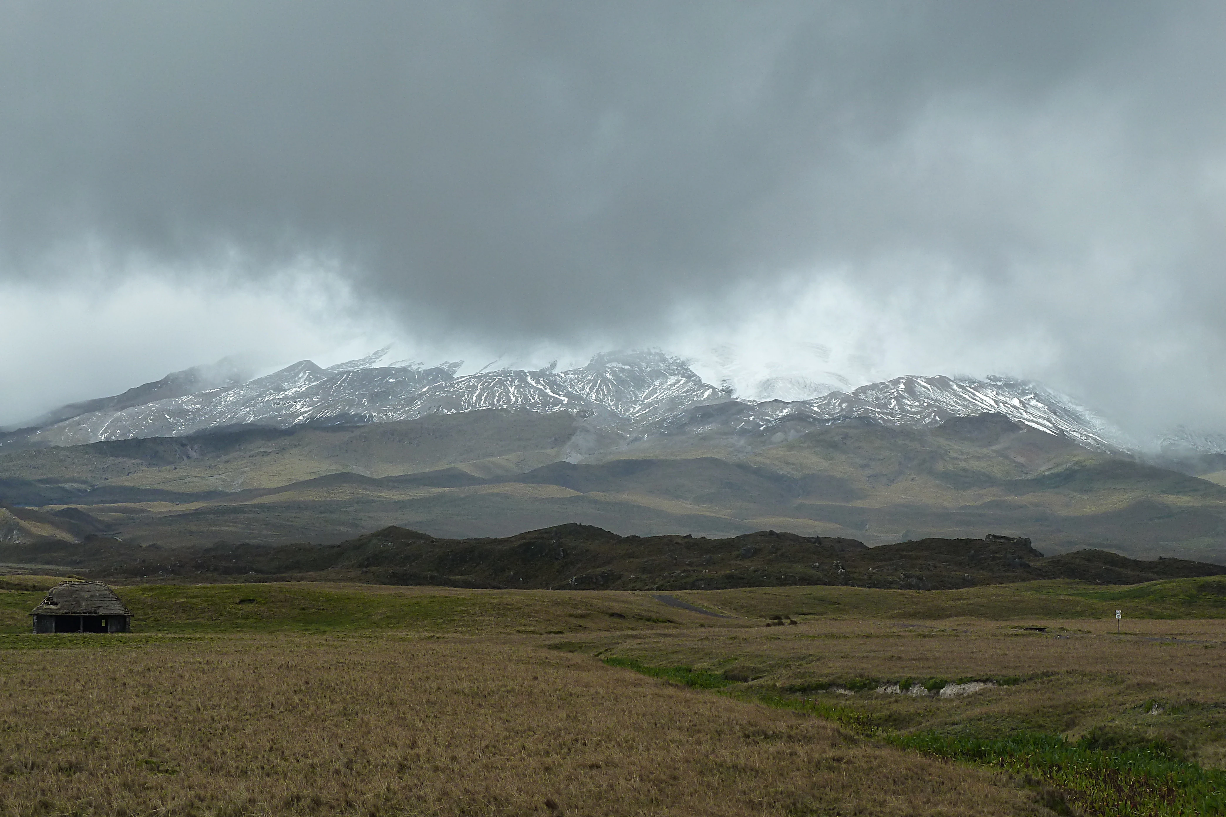 A wide shot of the snowy, rugged Antisana Volcano partially obscured by heavy grey clouds, overlooking a vast, rolling high-altitude grassland plain with a small structure visible in the lower left.