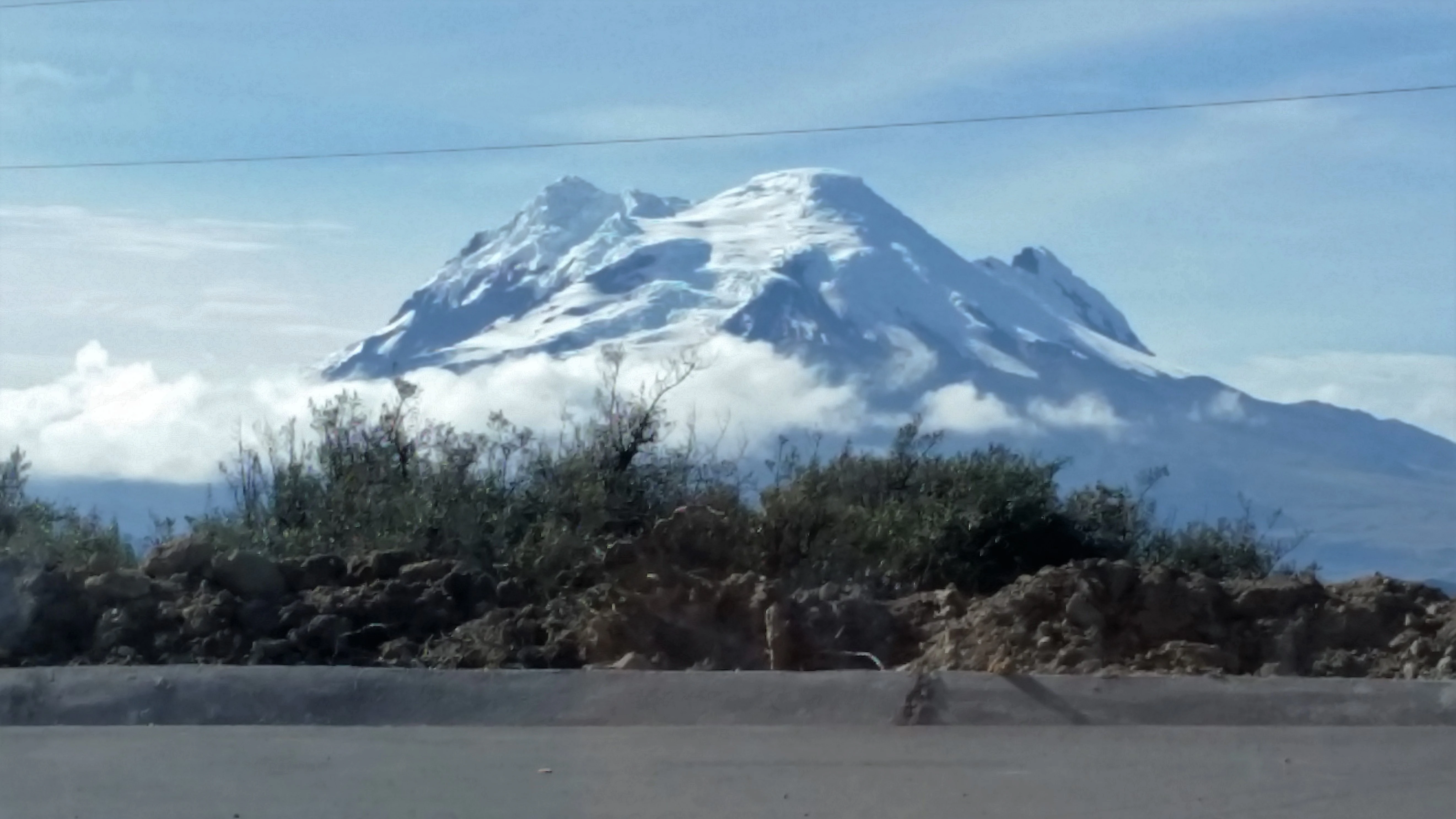 A wide shot of the Antisana volcano with a snow-capped summit partially obscured by clouds, viewed over a roadside area with rocky, scrubby vegetation in the foreground.