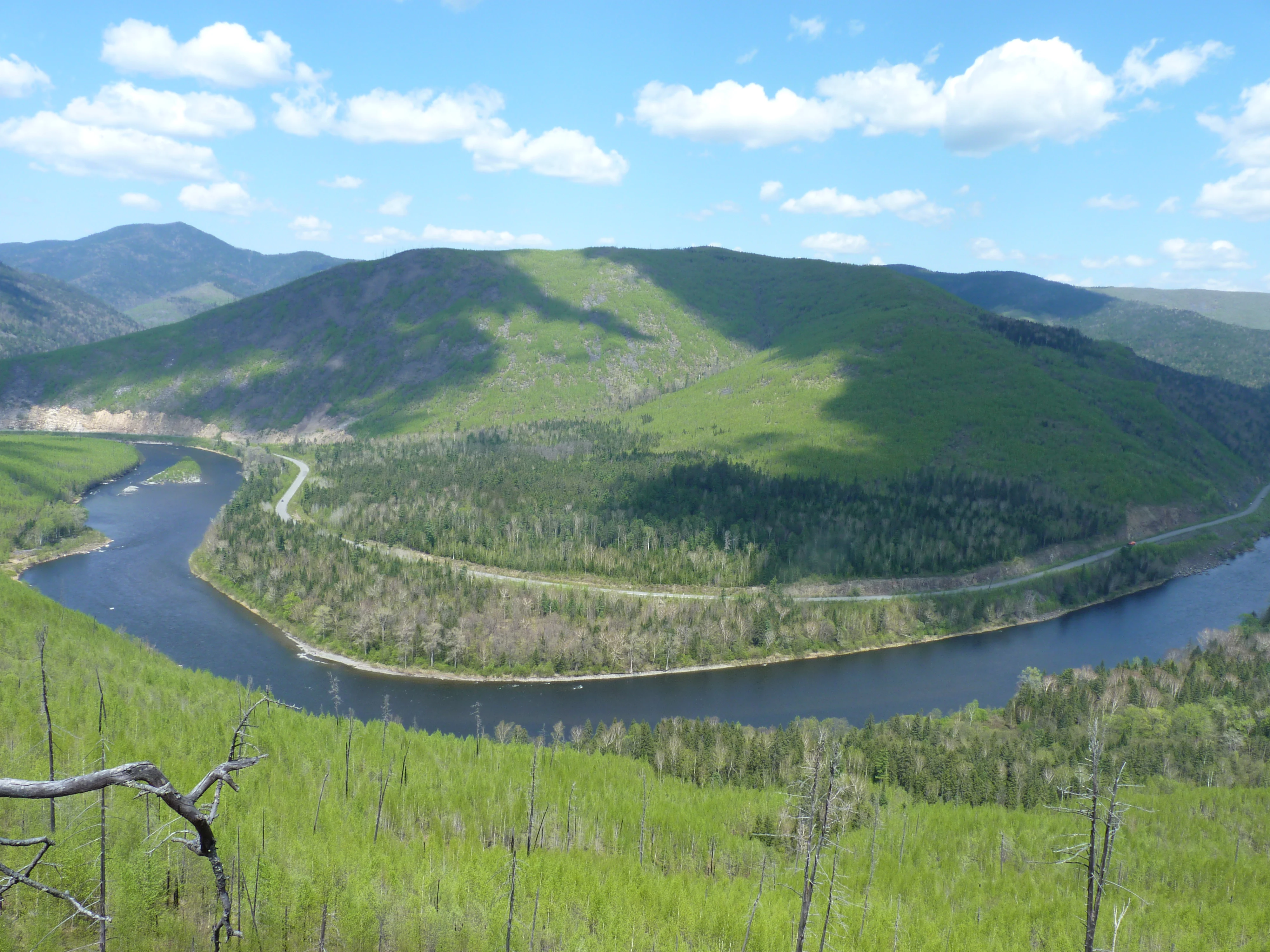 A wide-angle landscape photograph showing a river winding through a valley surrounded by green, forested mountain slopes under a partly cloudy blue sky.