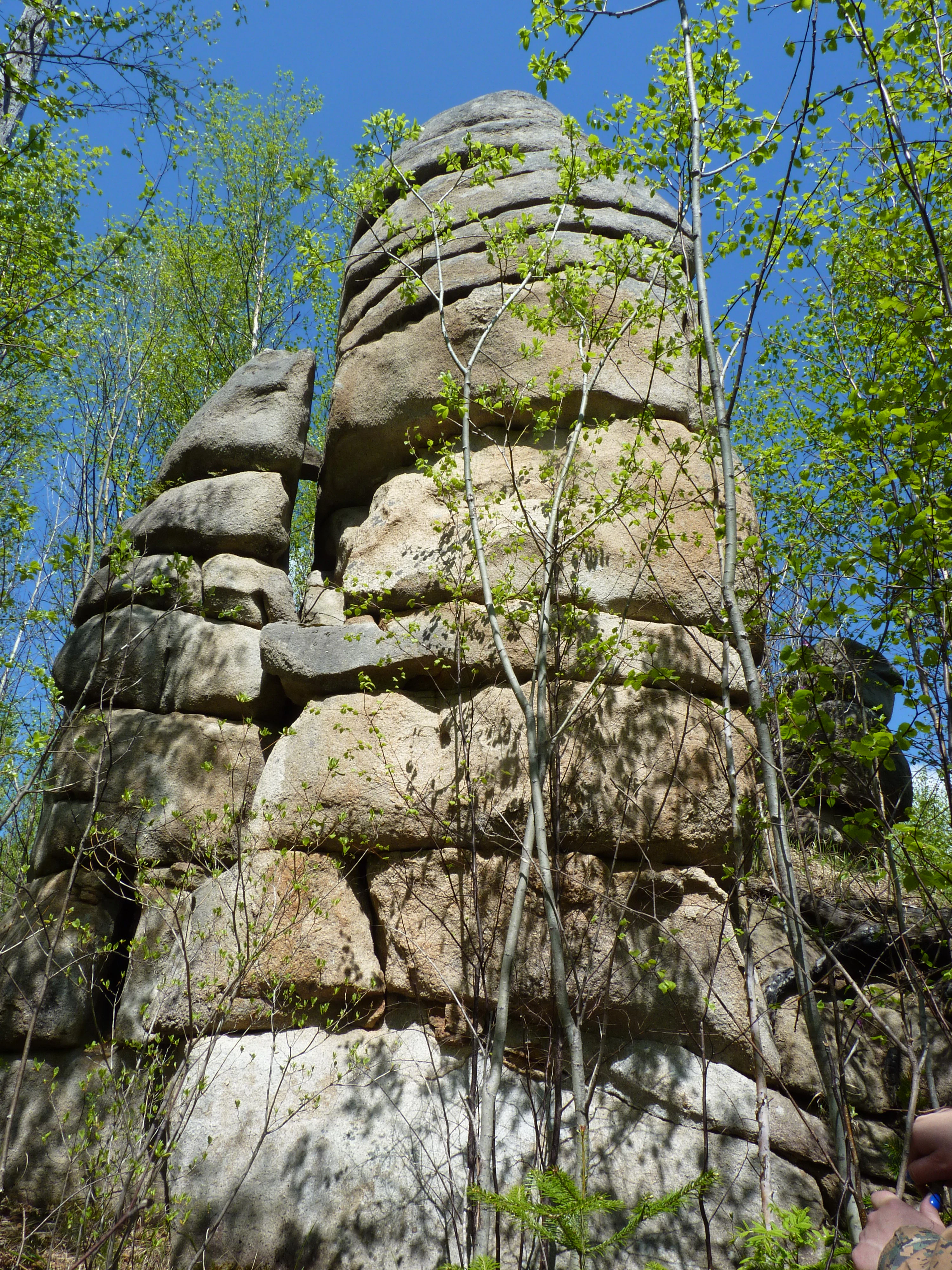 A large, vertically layered rock pillar formation surrounded by green deciduous trees under a clear blue sky.