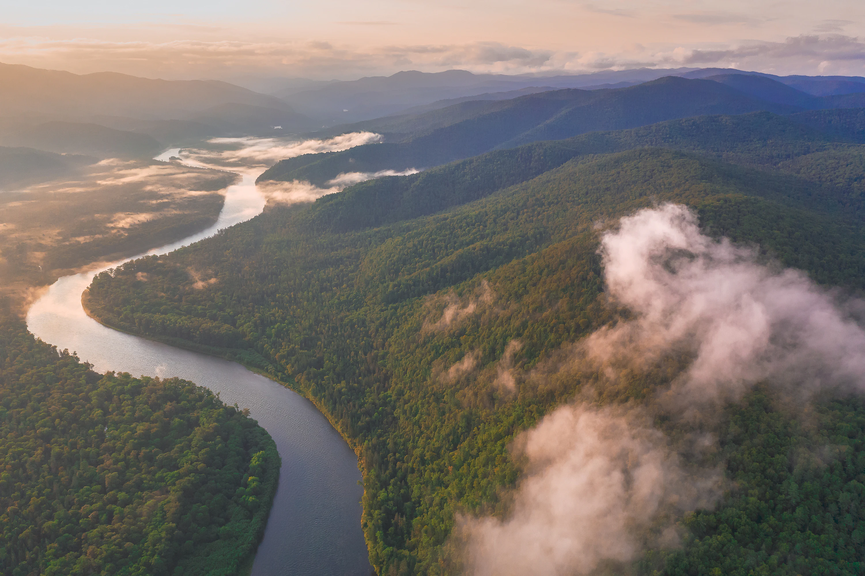 An aerial view of a winding river flowing through a lush, forested valley at sunrise with low-hanging clouds and mountain ridges in the distance.