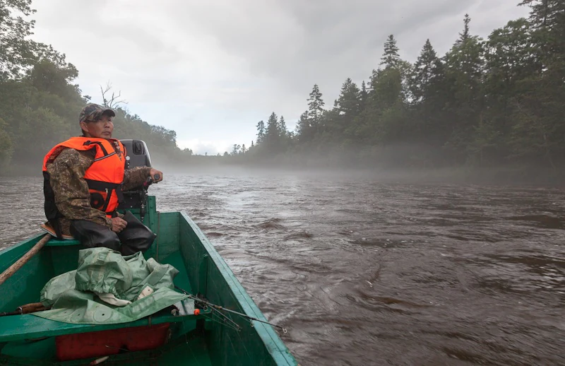 A person wearing a camouflage jacket and a bright orange life vest sits in a green motorboat on a river, with misty, tree-covered riverbanks in the background.