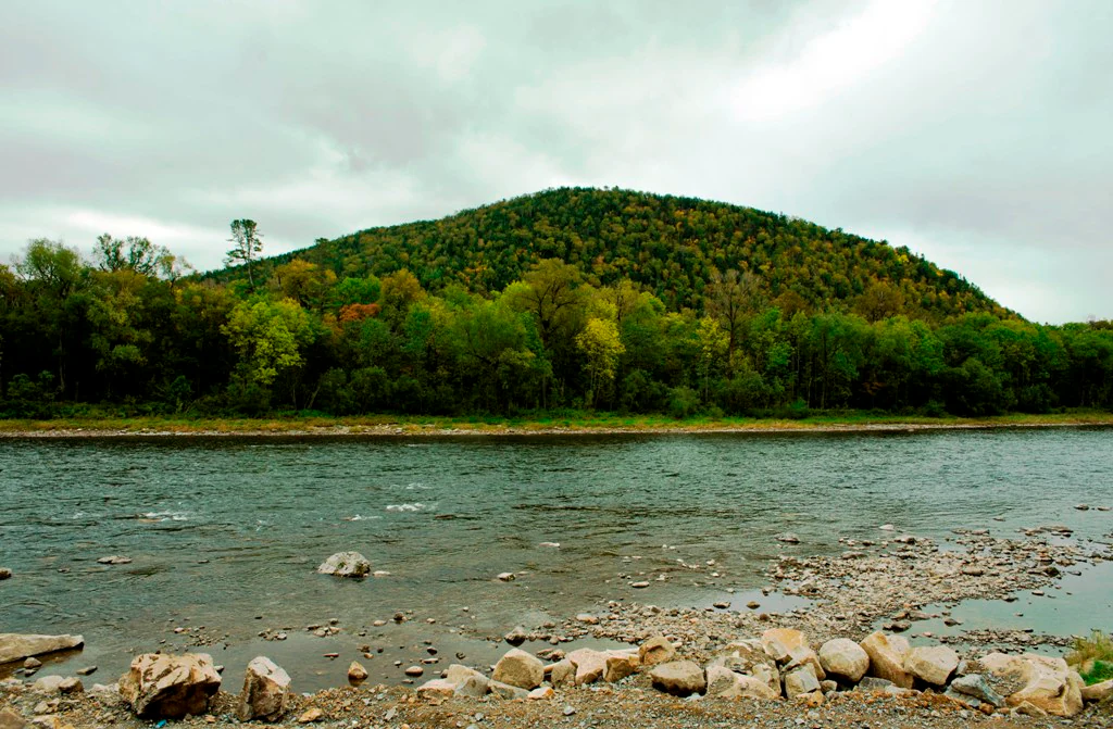 A wide view of the Anyuy River flowing in front of a forested, rounded mountain hill under an overcast sky.