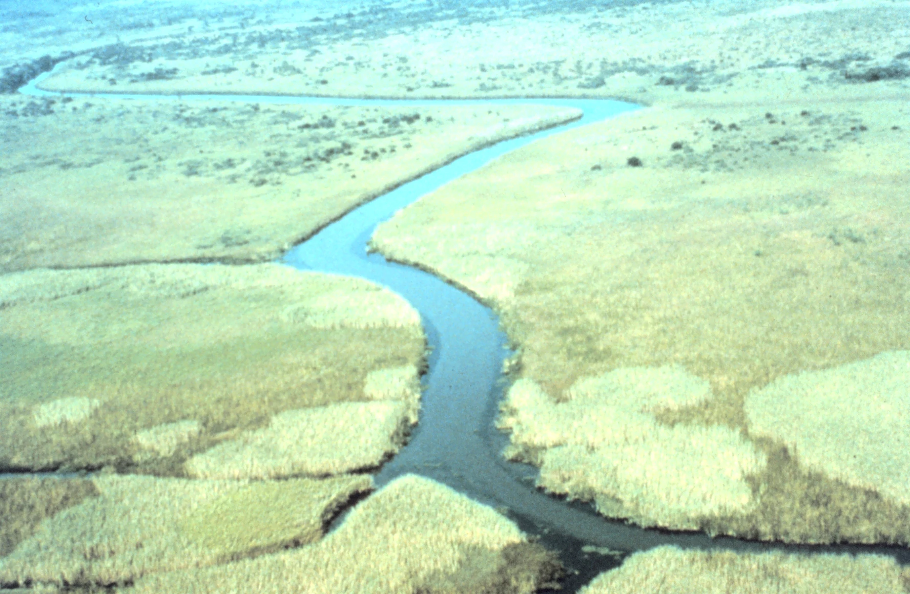 Una vista aérea en ángulo alto de un canal fluvial serpenteante que atraviesa extensas marismas y humedales.