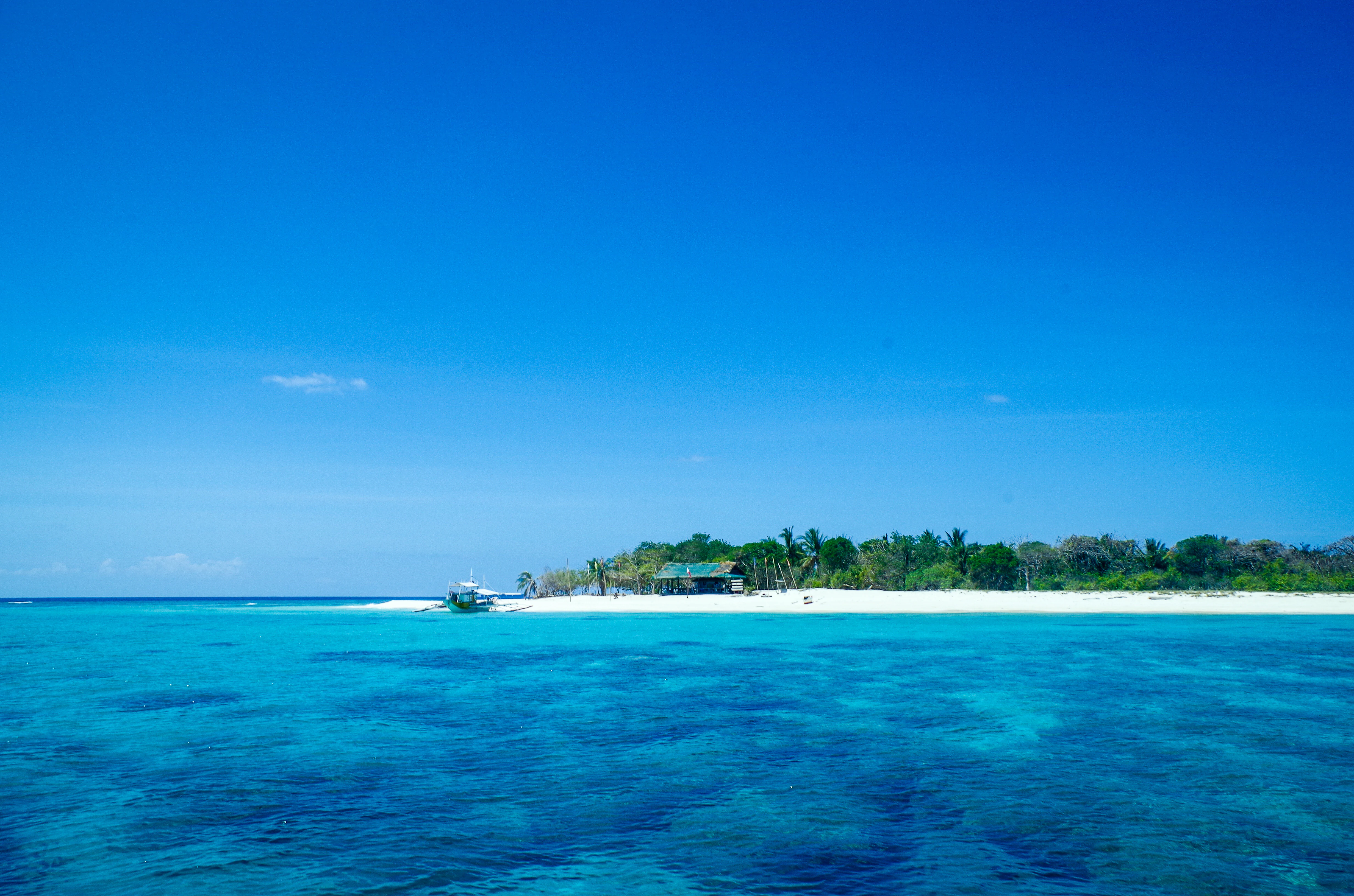 A tropical island with a white sandy beach and green trees surrounded by clear blue and turquoise water under a bright blue sky.