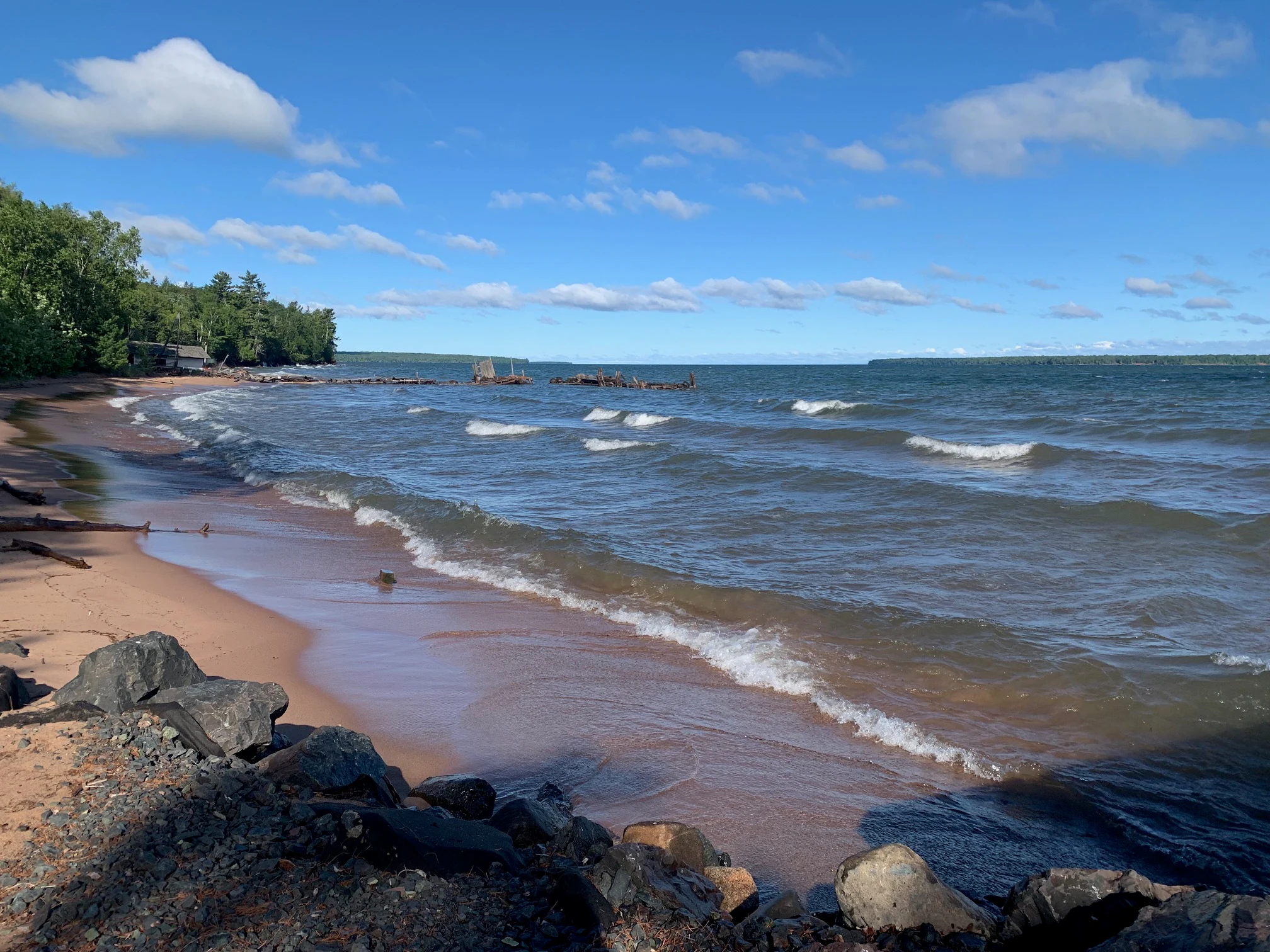 A sandy beach with small waves rolling in, bordered by a forest on the left and rocks in the foreground, under a blue sky with scattered clouds.