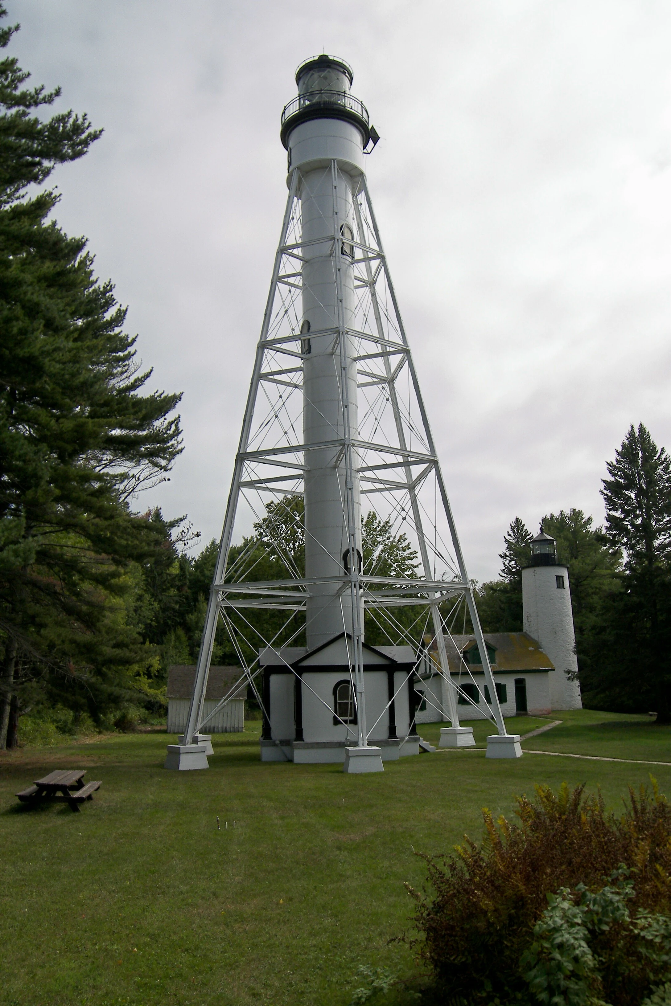 A tall white skeletal steel lighthouse tower stands on a grassy lawn with a shorter solid white lighthouse tower and small buildings in the background, surrounded by pine trees under an overcast sky.