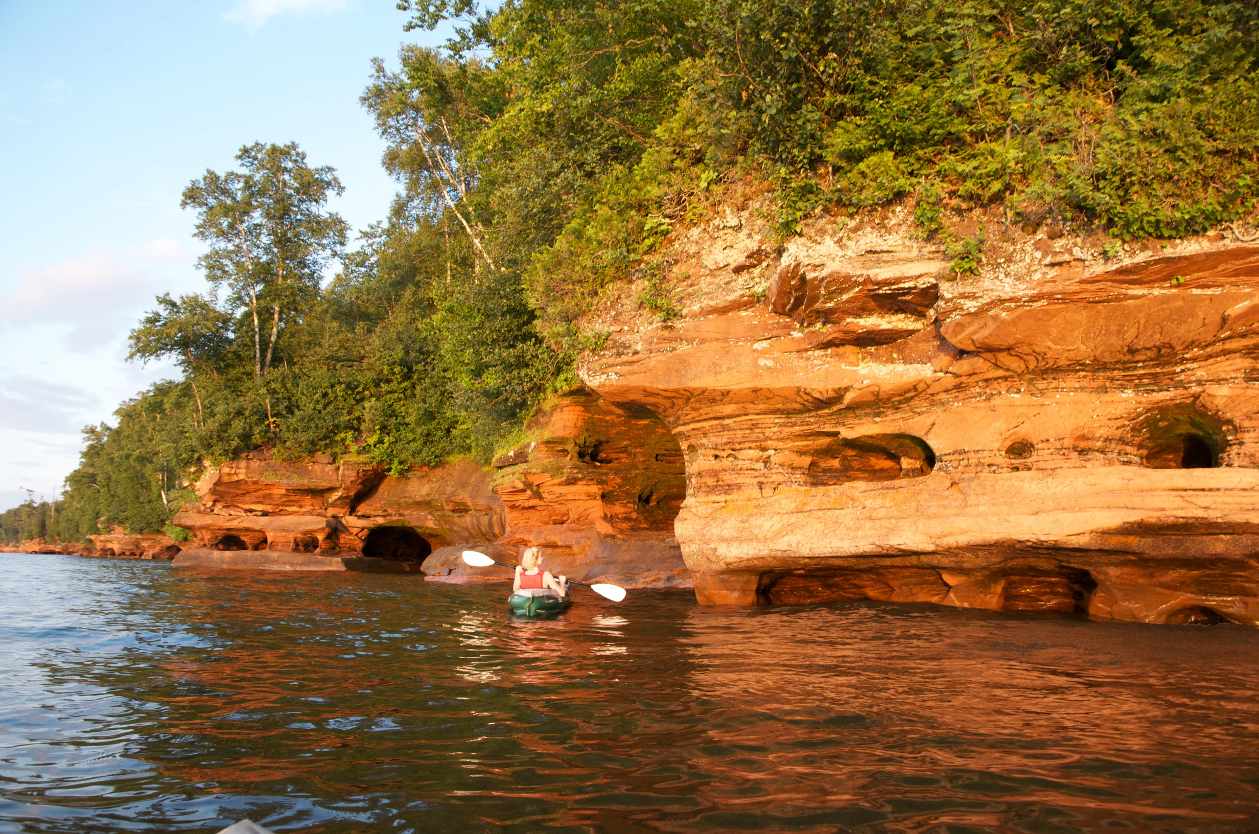 Two sea kayakers paddling on calm water near large red sandstone cliffs with sea caves and green trees on top.